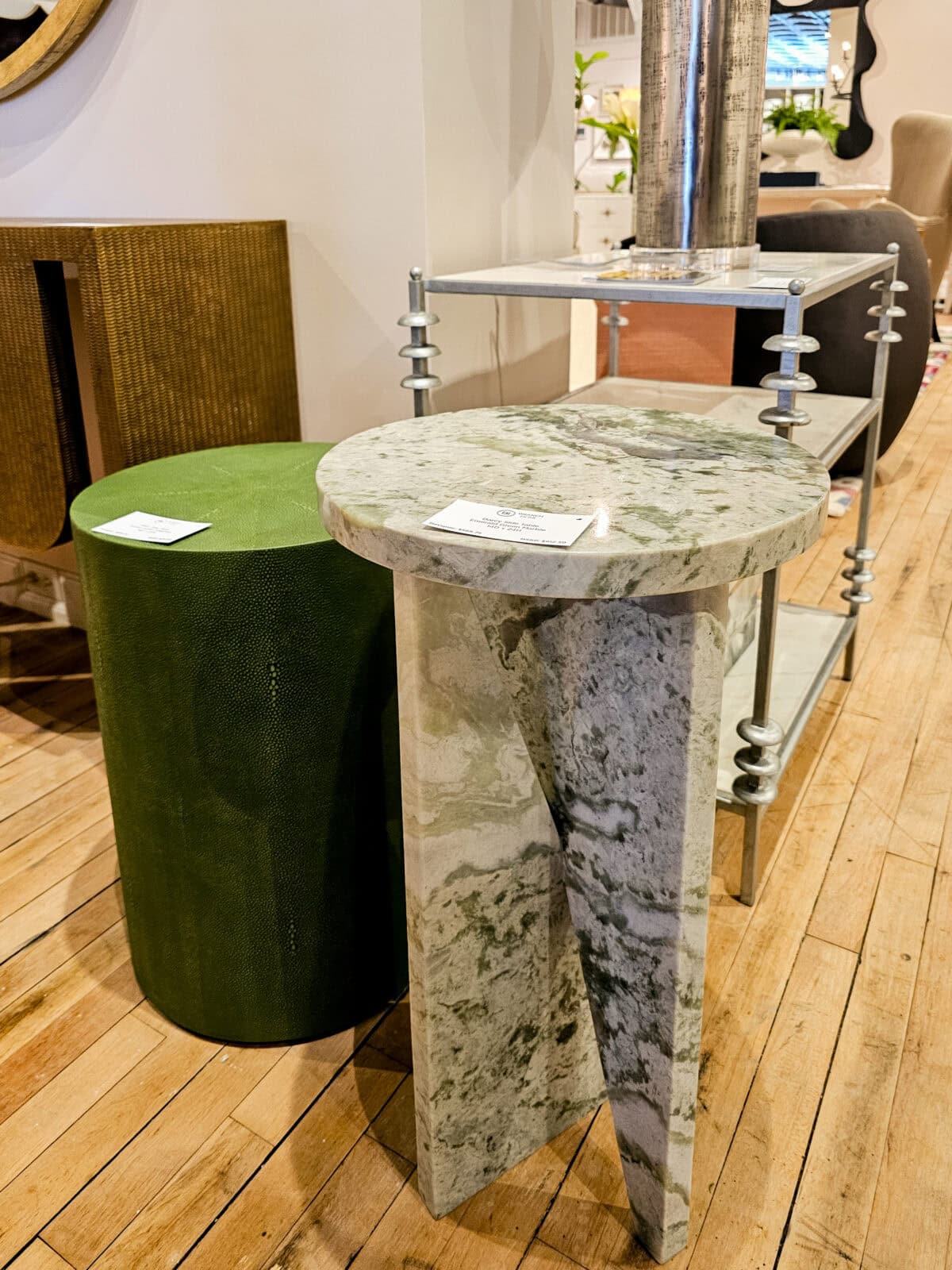 A green cylindrical table, a marble round-top pedestal table, and a metal-frame shelving unit are displayed on a wooden floor in a furniture store.