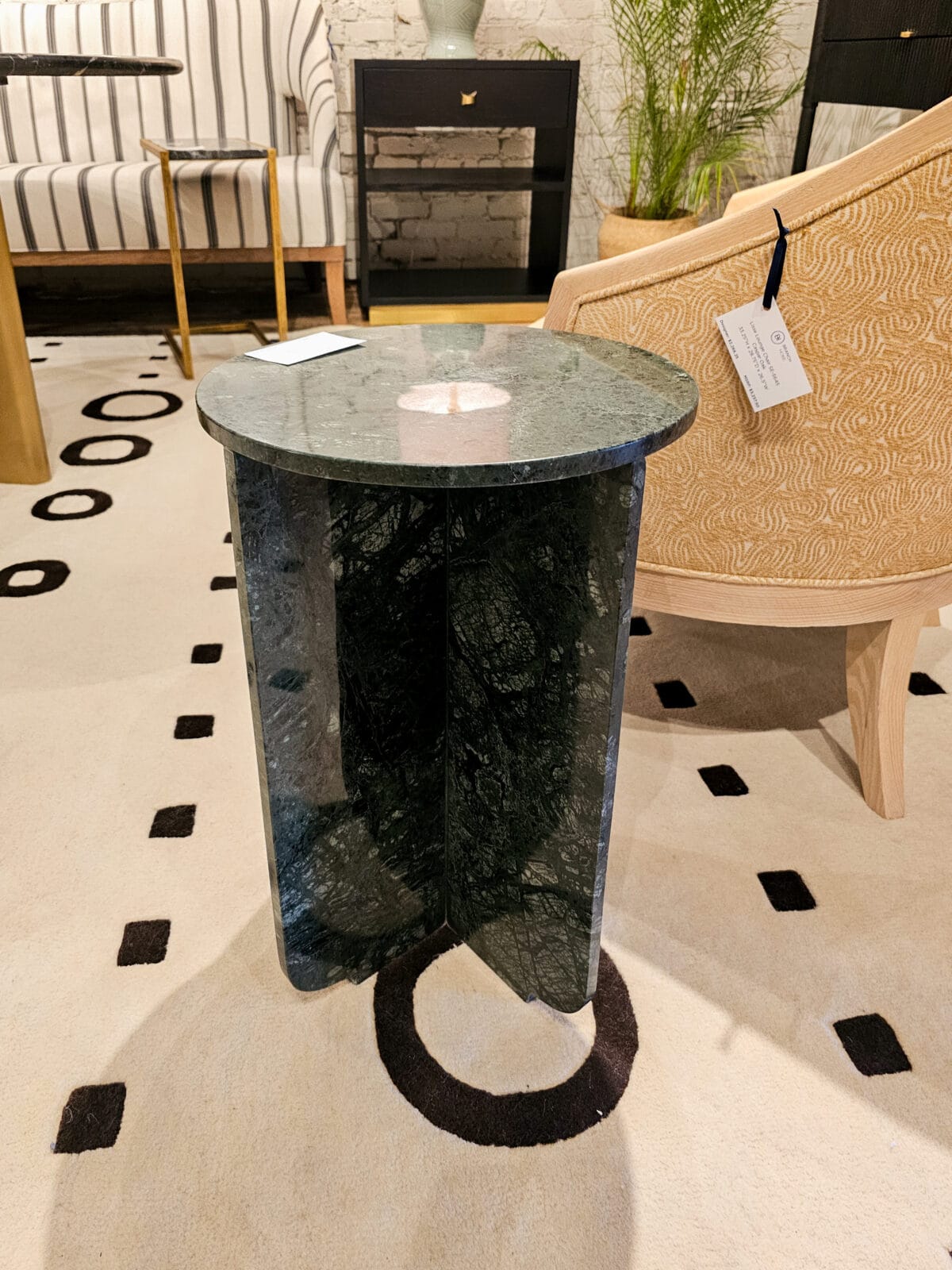 A small round green marble side table with cross-shaped base stands on a cream rug with black geometric patterns, surrounded by modern furniture.