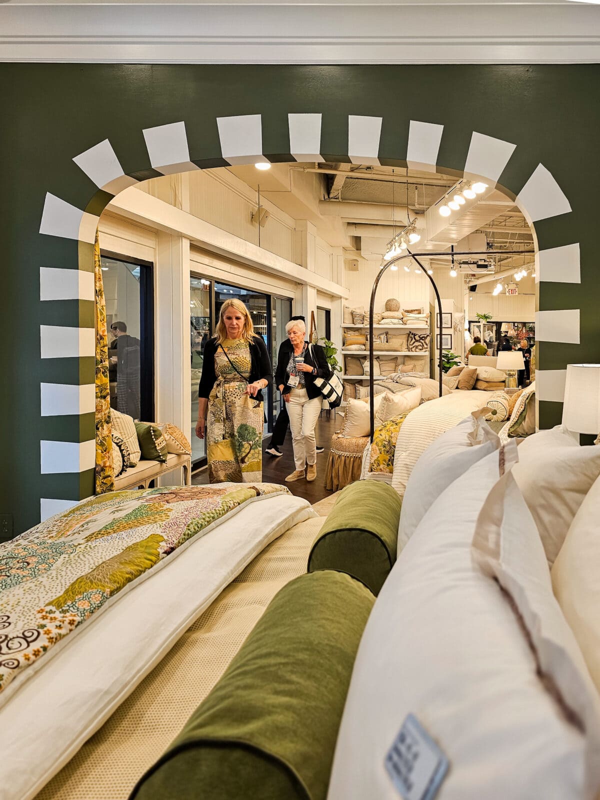 Two women walk through an arched doorway into a bedding store, surrounded by neatly arranged pillows, comforters, and home decor items.