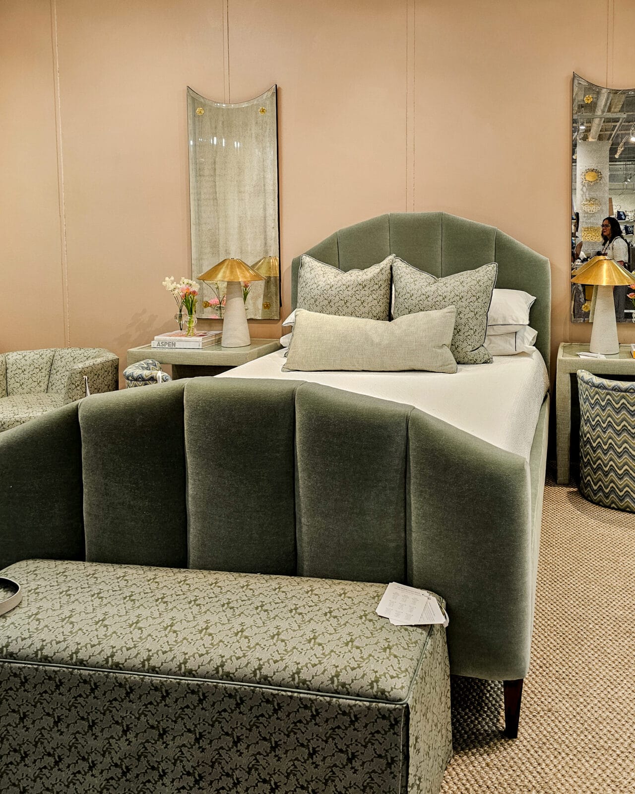 A bedroom with a green upholstered bed, matching bench, patterned pillows, two side tables with lamps, and two mirrors on a beige wall.
