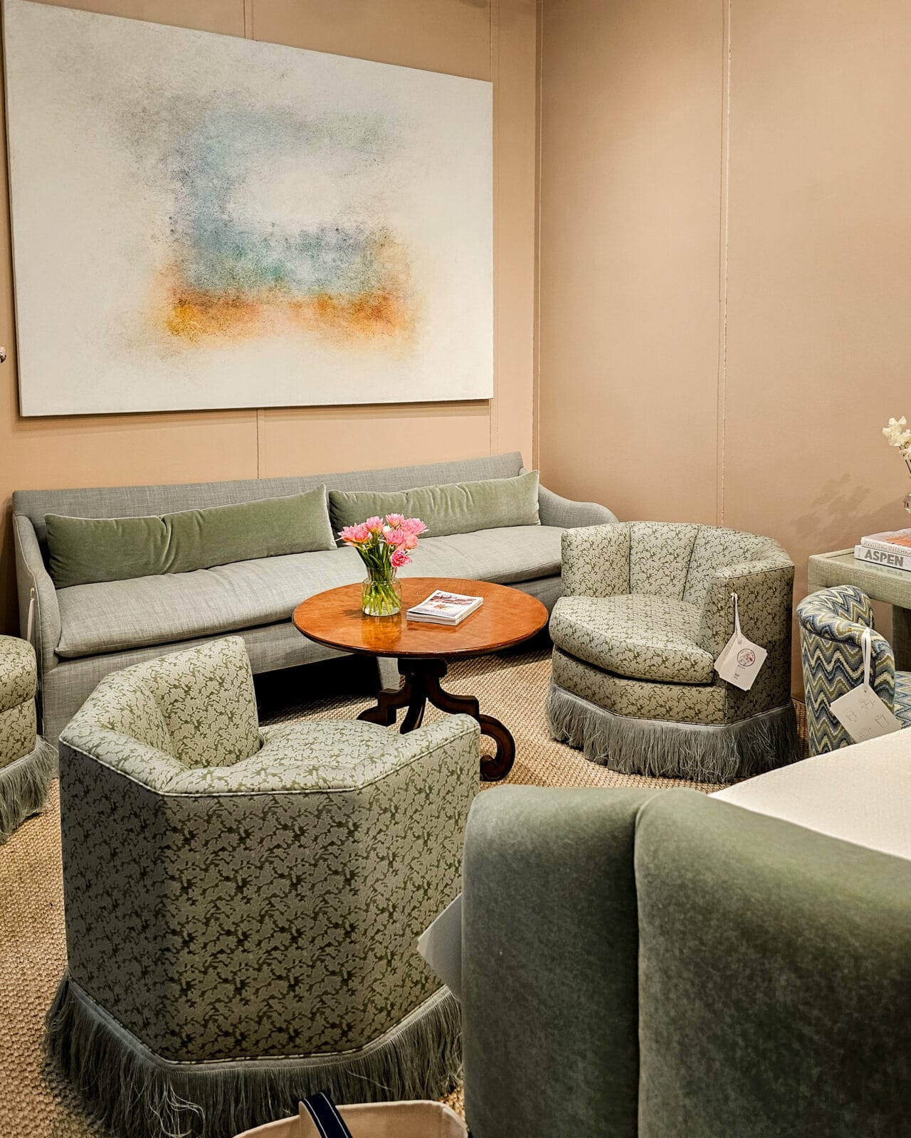 A living room with light green upholstered furniture, a round wooden coffee table with a vase of pink flowers, and an abstract painting on a beige wall.