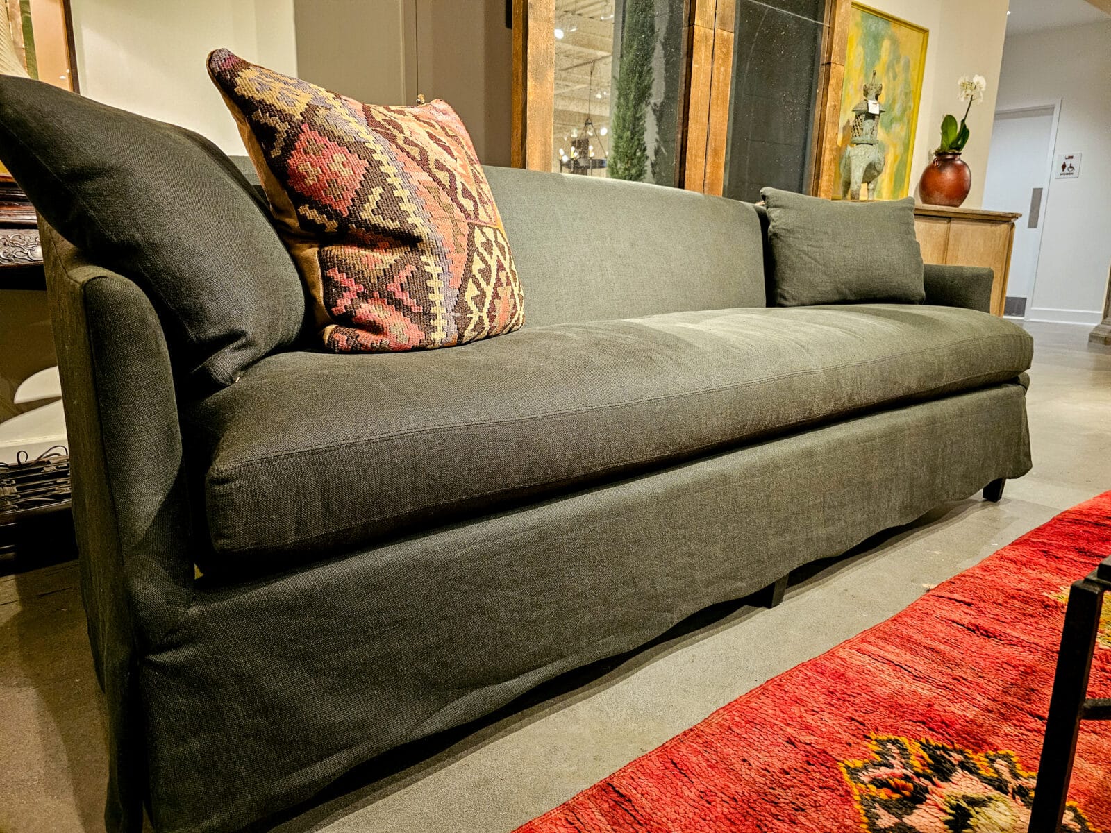 A dark gray upholstered sofa with three cushions, a patterned pillow, and a red rug placed in front, set in a room with wooden furniture and decor.