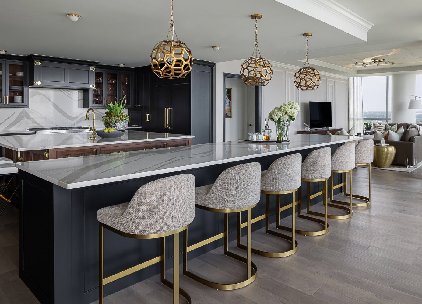 Modern kitchen with a large marble island, five upholstered bar stools with gold frames, pendant lights, and black cabinetry. Open layout with living room in the background.