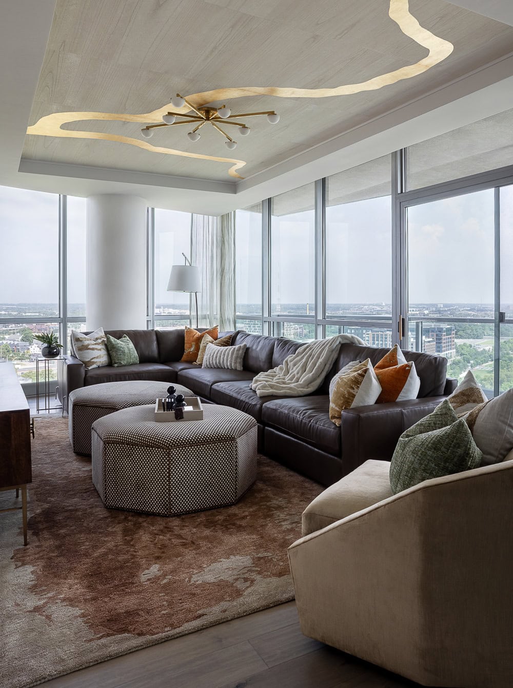 Modern living room with a large sectional sofa, patterned ottomans, accent pillows, floor-to-ceiling windows, city view, and a sculptural ceiling light fixture.