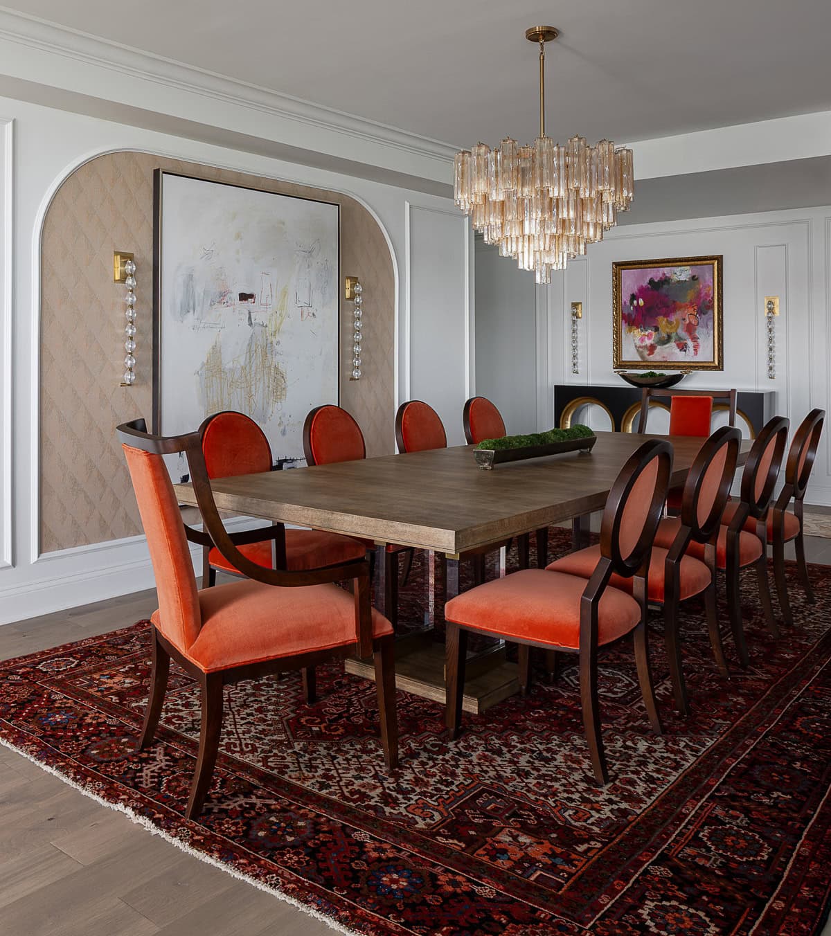 A dining room with a wooden table, eight red-cushioned chairs, a patterned rug, a modern chandelier, and abstract artwork on white and beige paneled walls.