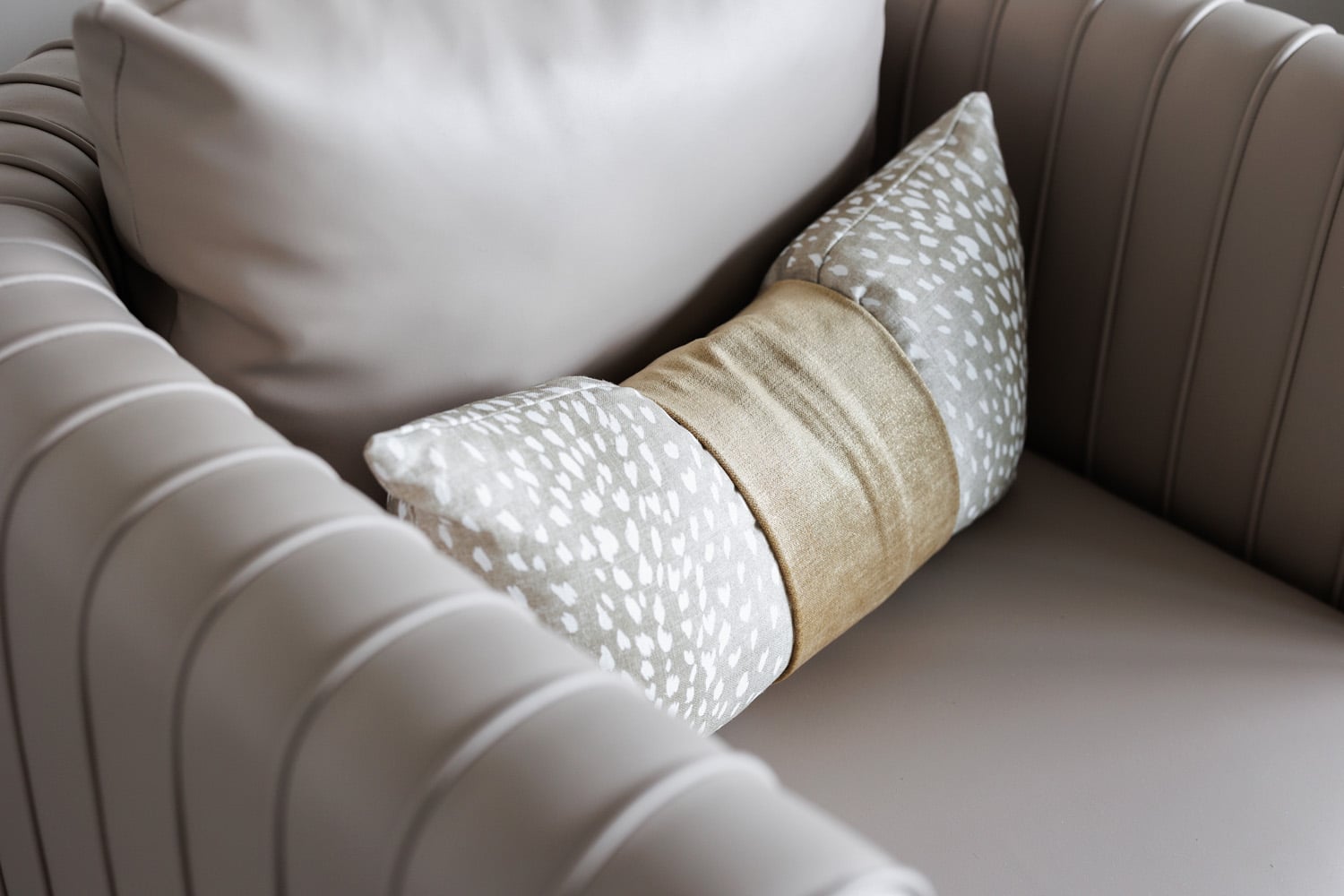 Beige and gray pillow with a gold center placed on a light gray, ribbed upholstered armchair.