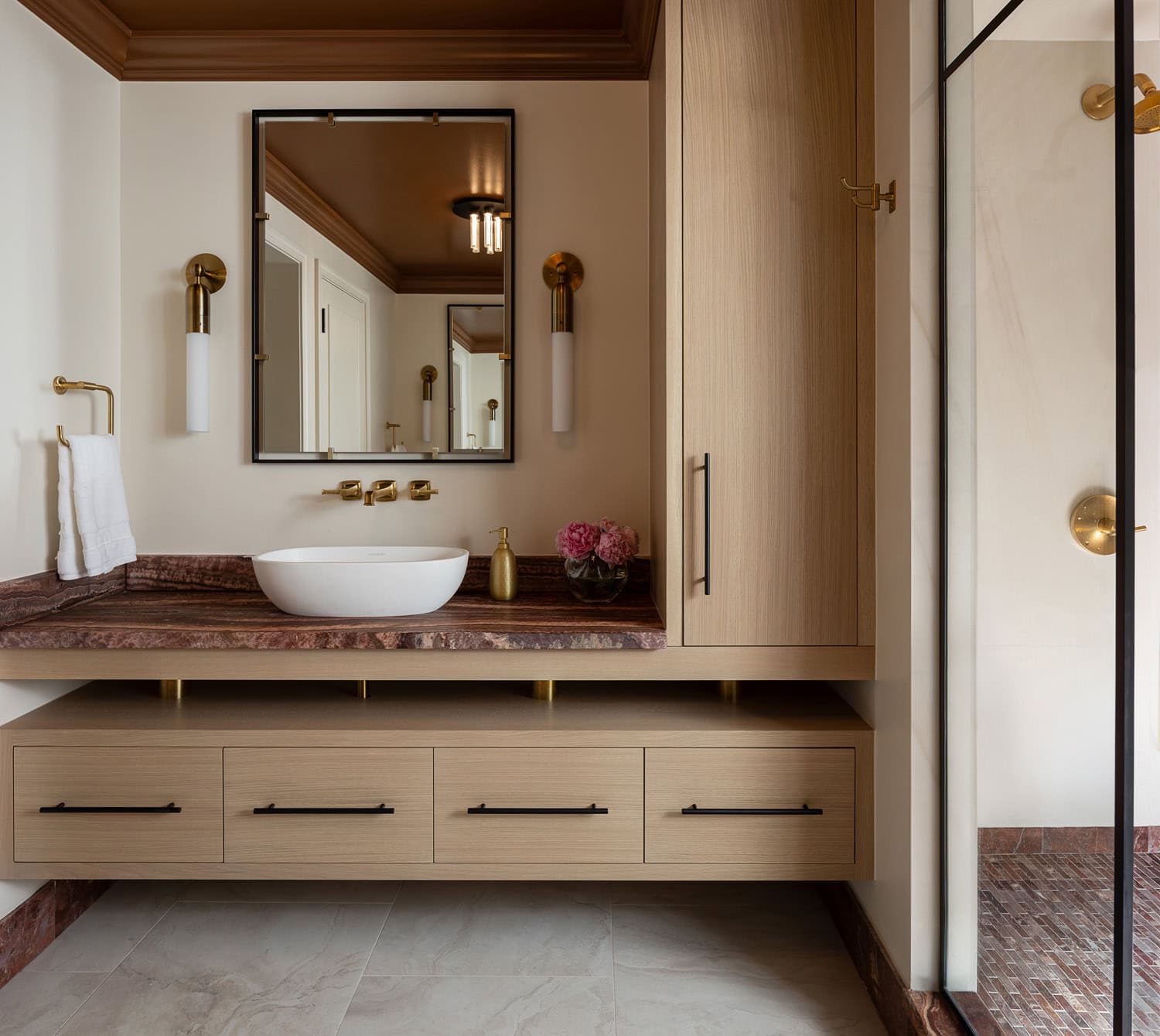 Modern bathroom with a floating wood vanity, vessel sink, wall-mounted faucet, large mirror, gold fixtures, and a glass shower enclosure. Pink flowers sit next to the sink.