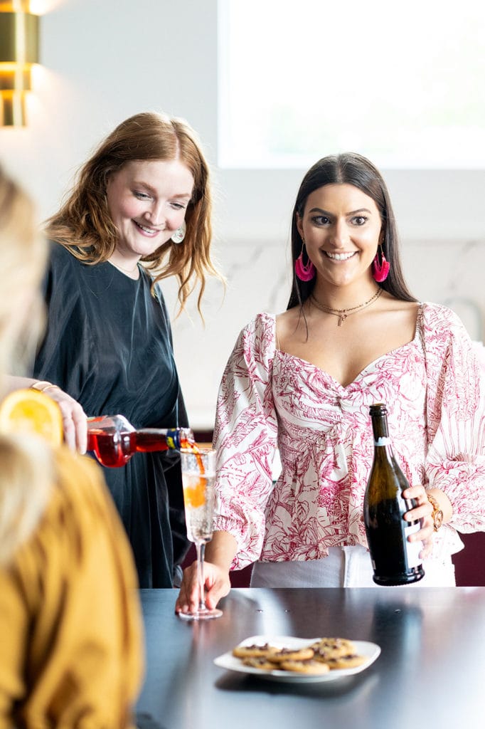 Two women stand at a table; one pours a drink into a glass while the other holds a bottle. A plate of cookies sits on the table in front of them.