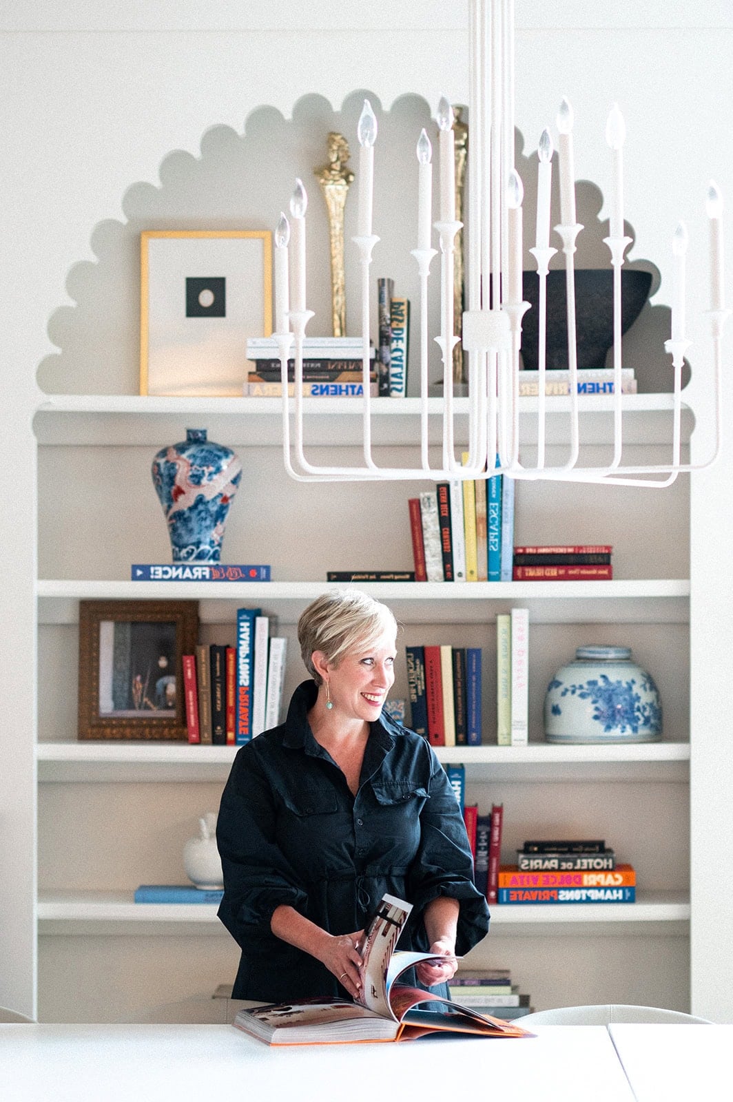 A woman stands at a table looking at an open book, in front of a built-in bookshelf filled with books, vases, and decorative items. A white chandelier hangs overhead.