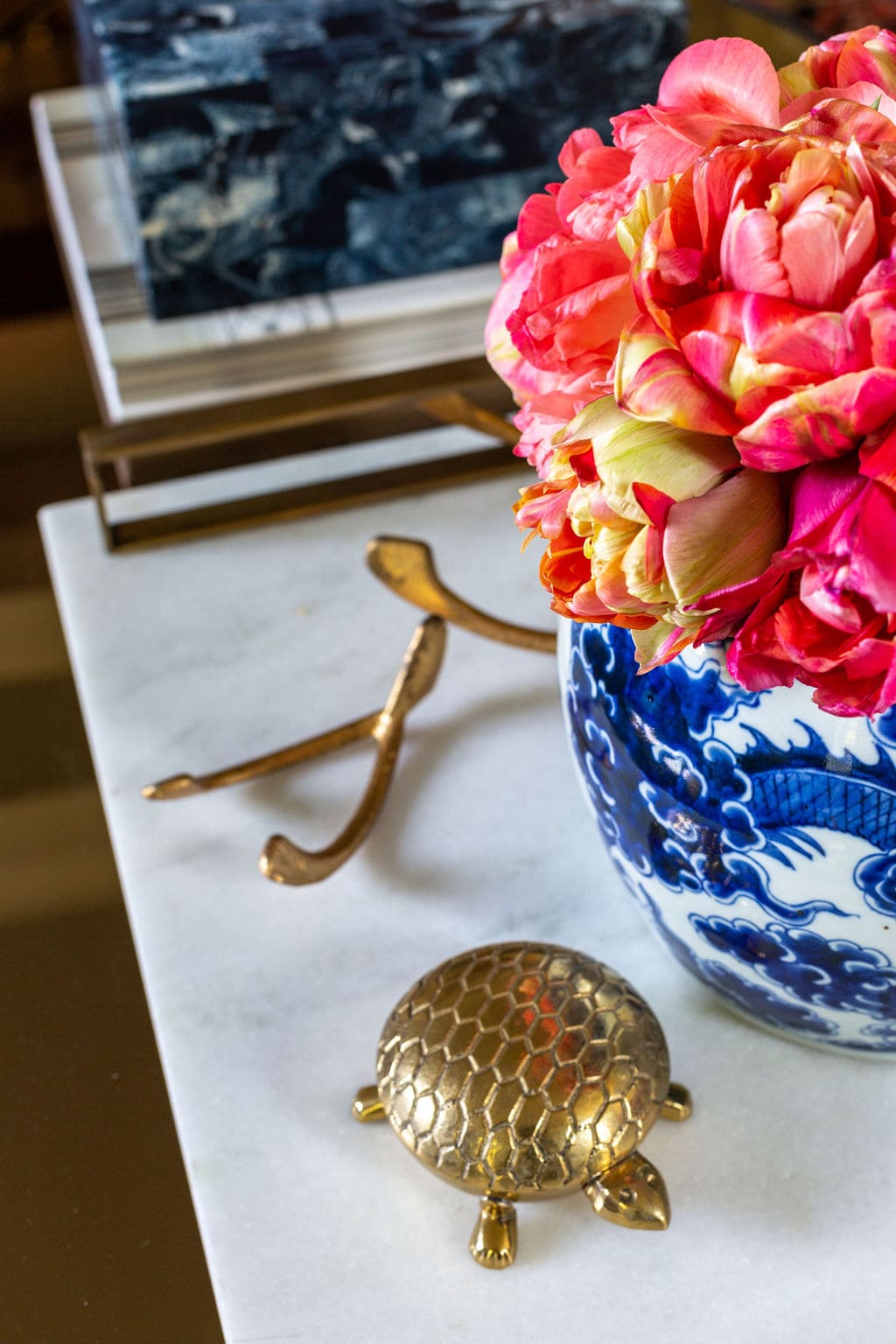 A blue and white vase with pink and yellow flowers sits on a marble surface next to a small brass turtle figurine and a decorative brass wishbone.