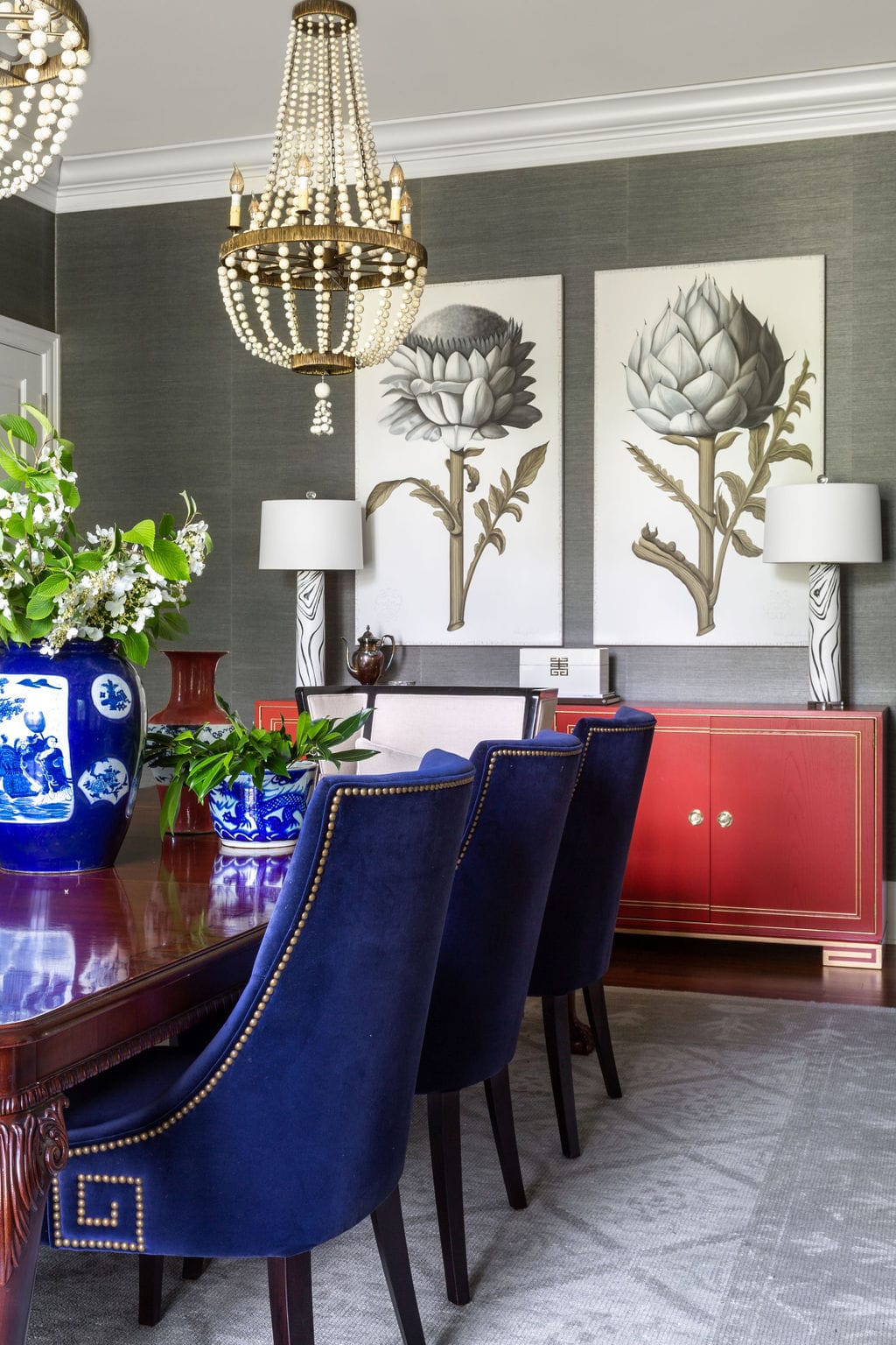 A dining room with blue velvet chairs, a wooden table, beaded chandeliers, floral wall art, a red sideboard, and blue-and-white vases with flowers.