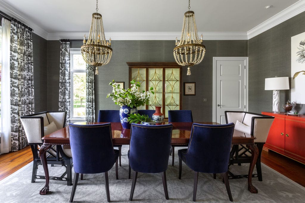 Formal dining room with a wooden table, eight blue velvet chairs, two chandeliers, a glass cabinet, red sideboard, and floral centerpiece on a patterned rug.