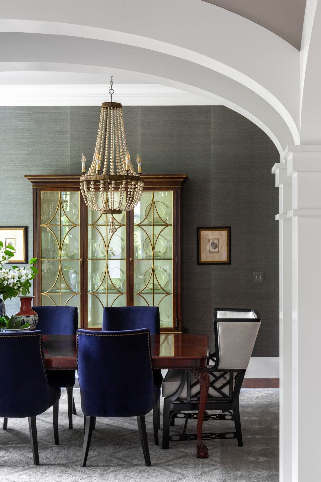 A formal dining room with a wood table, navy blue chairs, a pearl beaded chandelier, and a glass-front china cabinet against a dark gray wall.
