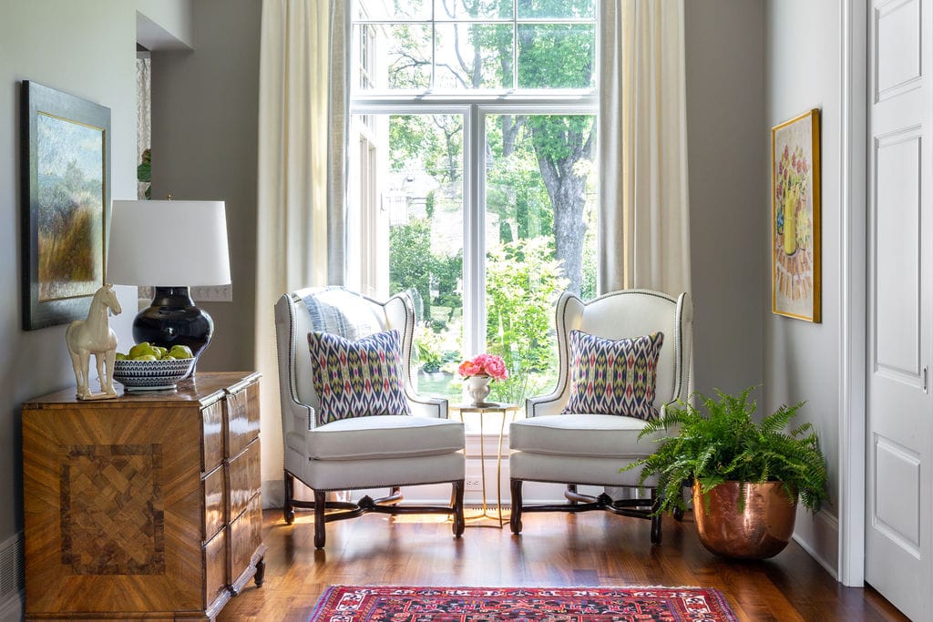 Bright sitting area with two white armchairs, patterned pillows, a side table with flowers, a chest with a lamp, and a large window with a garden view.