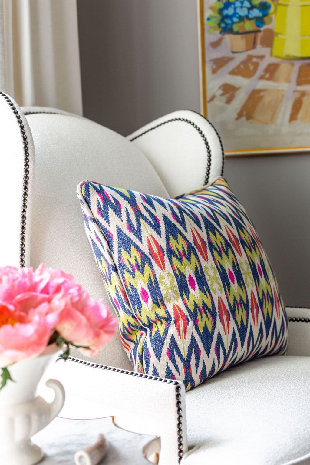 A white upholstered armchair with nailhead trim features a colorful patterned cushion; pink flowers and a framed painting are visible in the background.