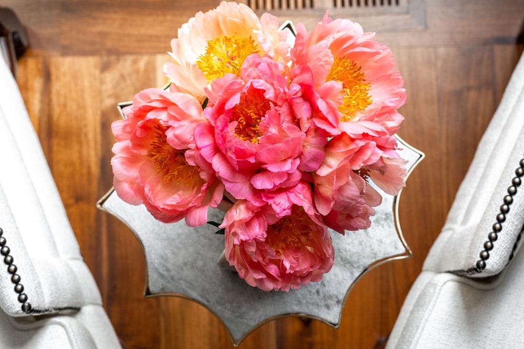 A bouquet of pink and peach peonies in a vase sits on a marble-top table between two white upholstered chairs on a wooden floor.