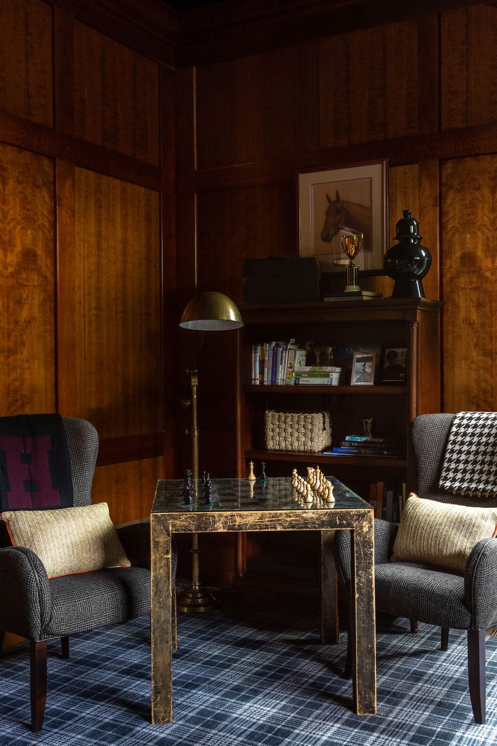 A chess table with pieces set up between two armchairs in a wood-paneled room with a bookshelf, lamp, and framed horse portrait.