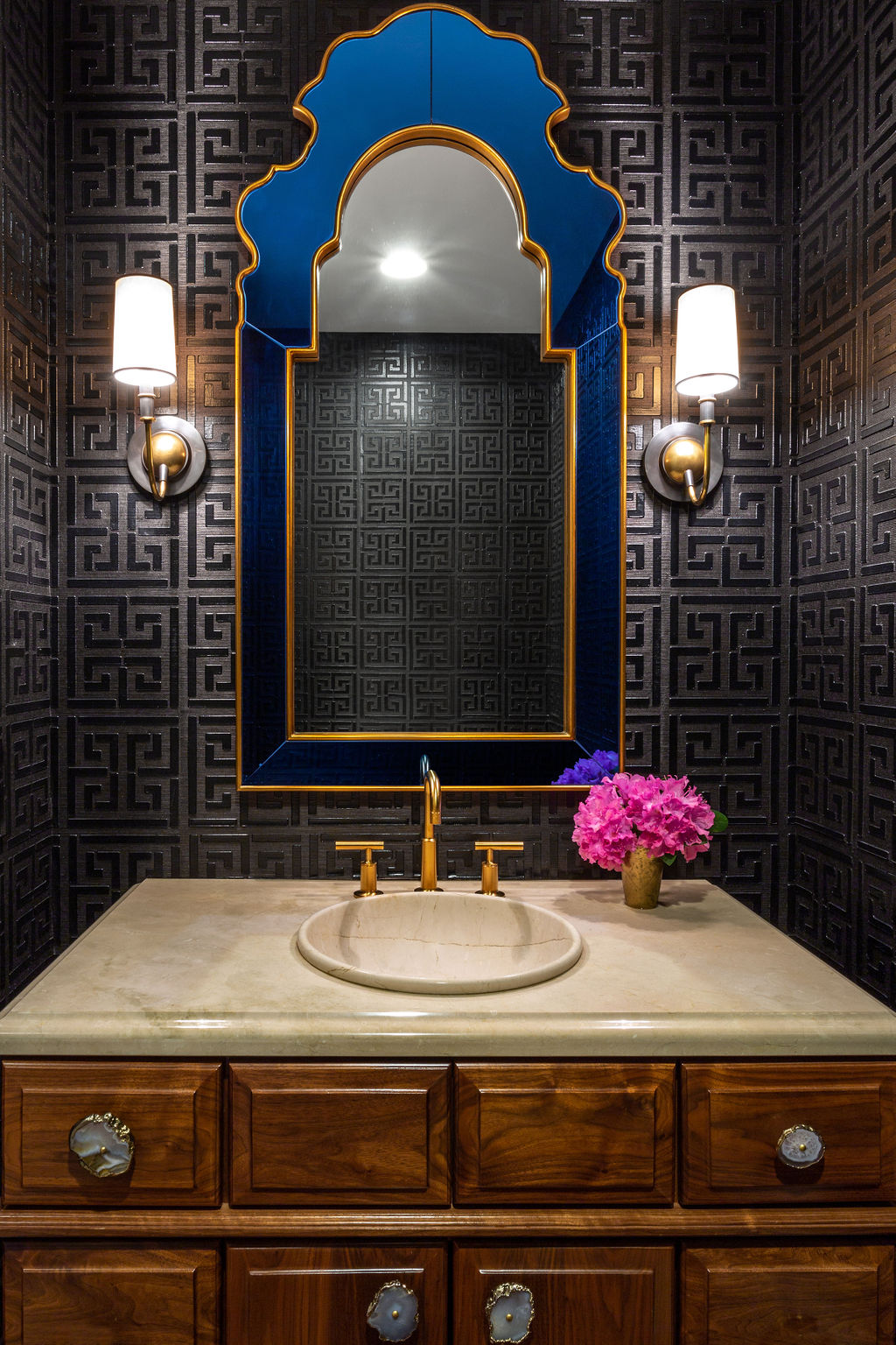 A bathroom vanity with a round sink, gold faucet, ornate blue mirror, two wall sconces, patterned dark wallpaper, and a small vase of pink flowers.