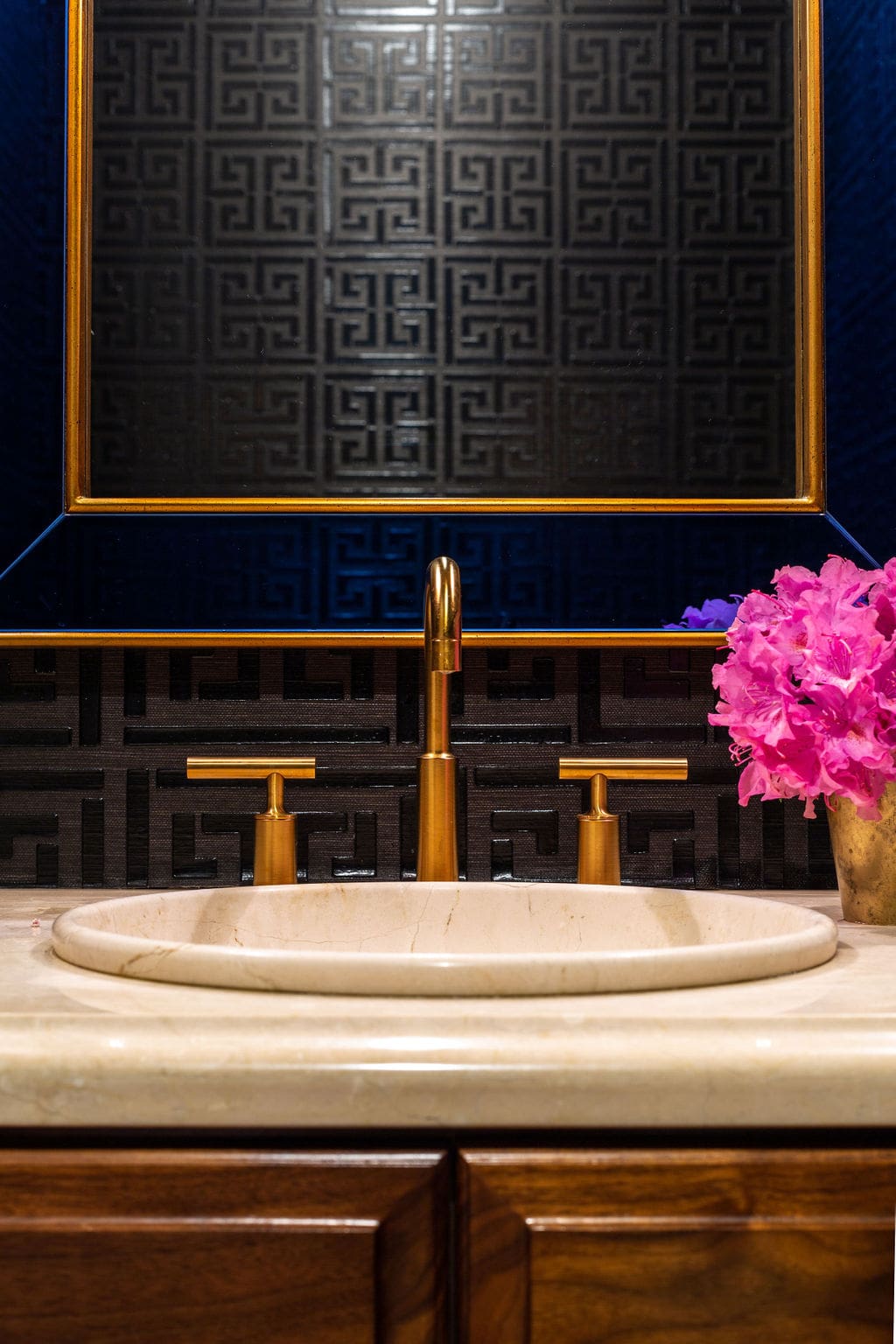 A bathroom sink with gold faucet and handles, beige countertop, dark geometric patterned wall, and a vase of pink flowers.