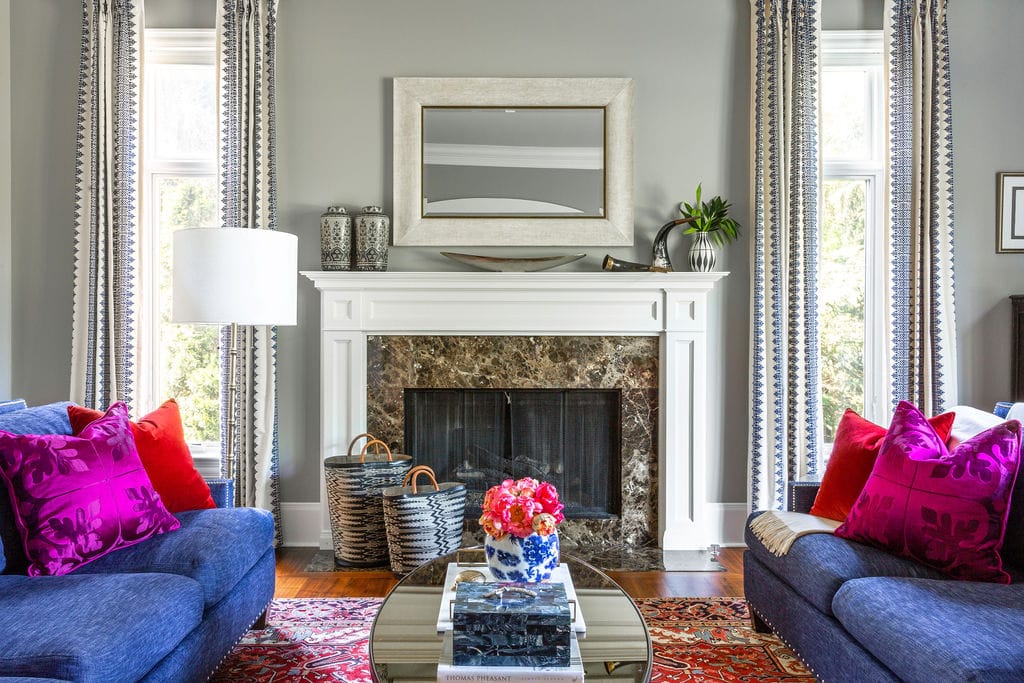Living room with a marble fireplace, framed mirror, blue sofas with pink pillows, red patterned rug, and a coffee table with books and a flower arrangement.