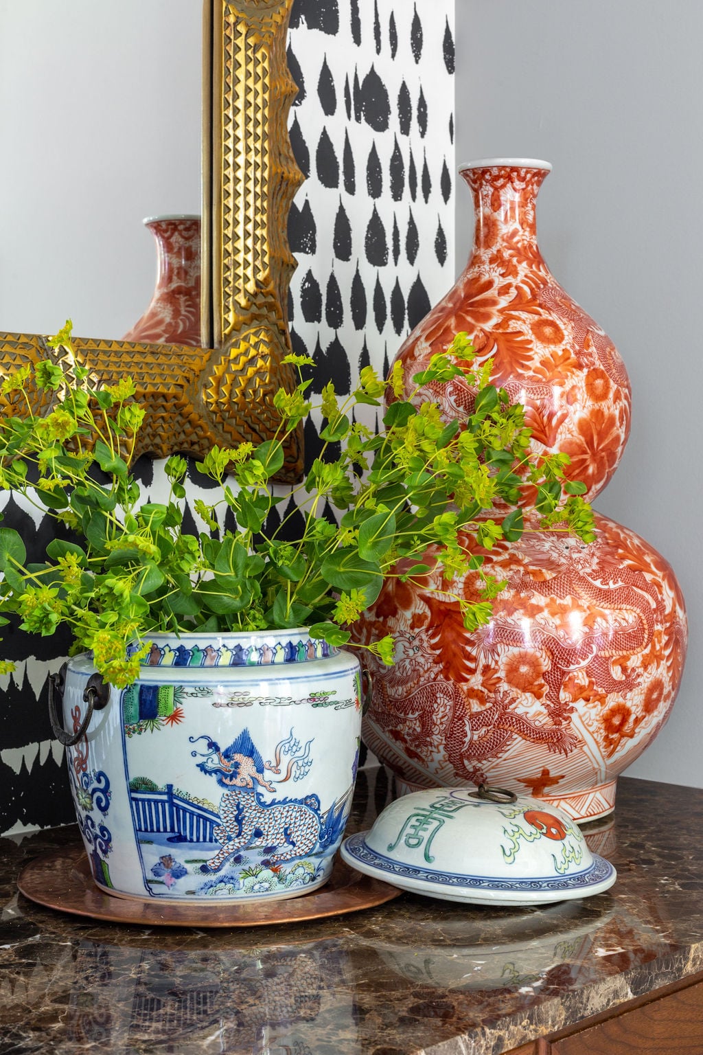 Blue and white ceramic pot with green foliage sits beside two tall red patterned vases and a gold-framed mirror on a marble tabletop.