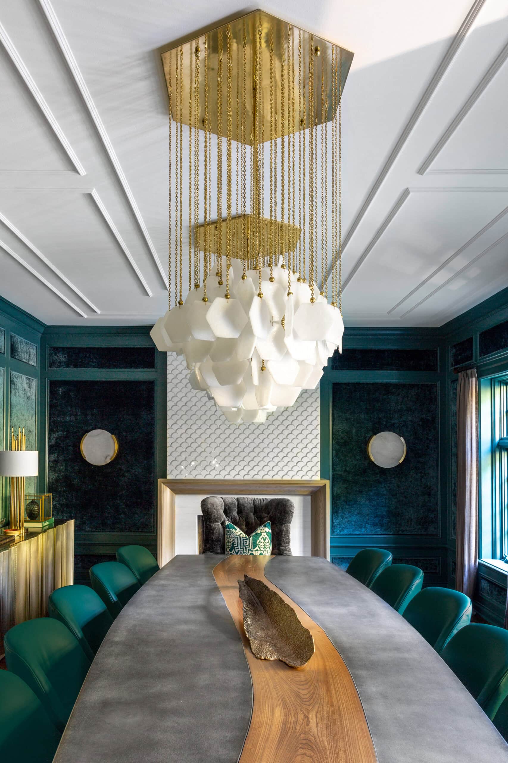 Luxurious dining room with a long grey table, teal chairs, a gold and white chandelier, dark blue walls, and geometric decor elements.