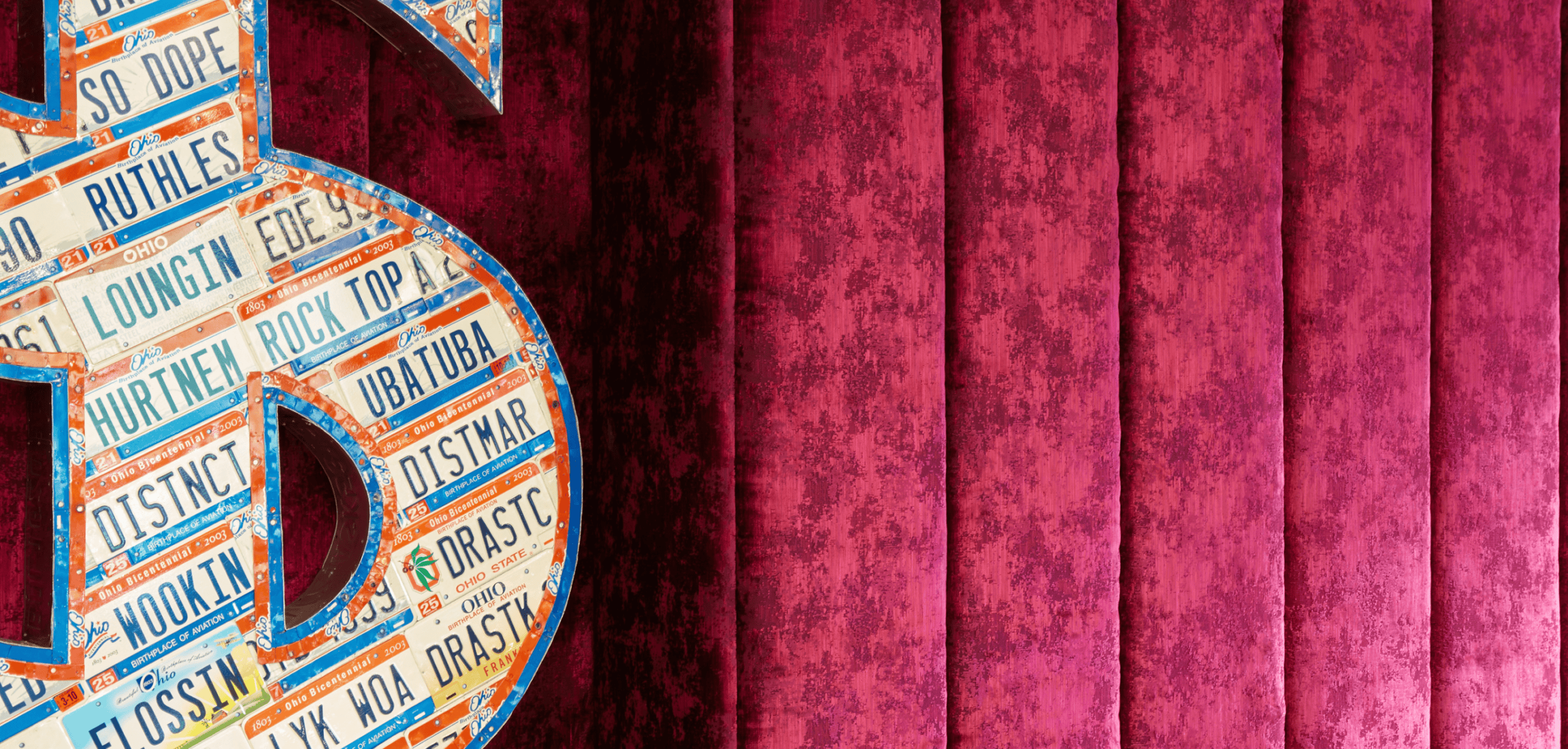 A large dollar sign made of colorful license plates stands beside a vertically paneled, textured red wall.