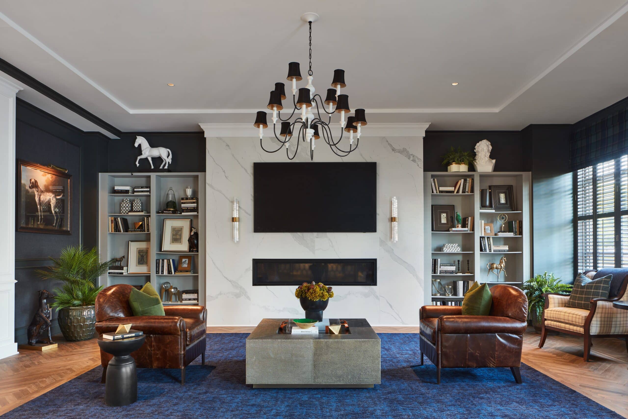 Modern living room with marble fireplace, wall-mounted TV, two brown leather chairs, a blue rug, bookshelves, a chandelier, and decorative items including a horse statue and framed art.