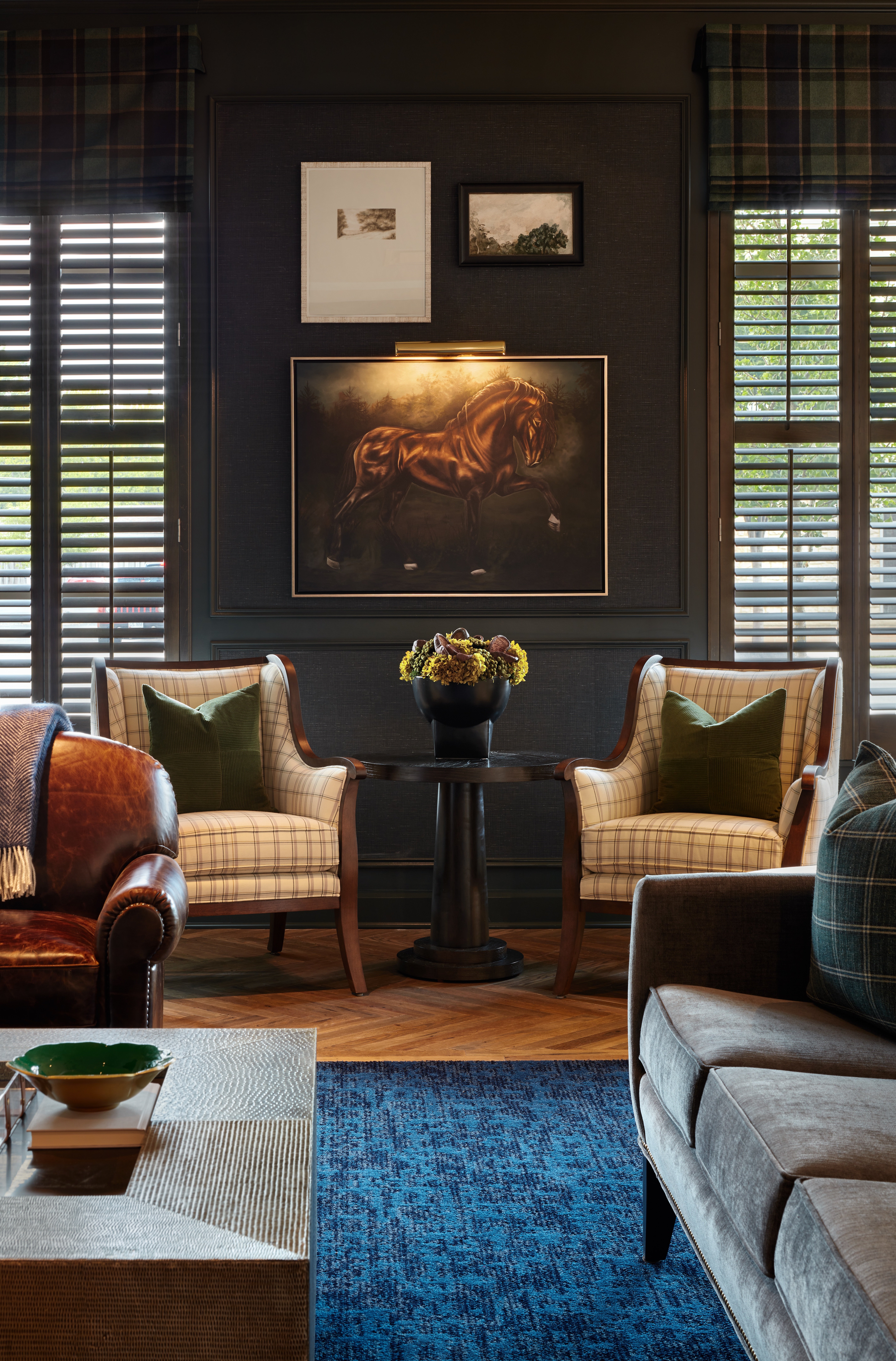 A living room by Crimson Design Group featuring two plaid armchairs, a small table with a potted plant, a horse painting, framed artwork, and large windows with shutters.