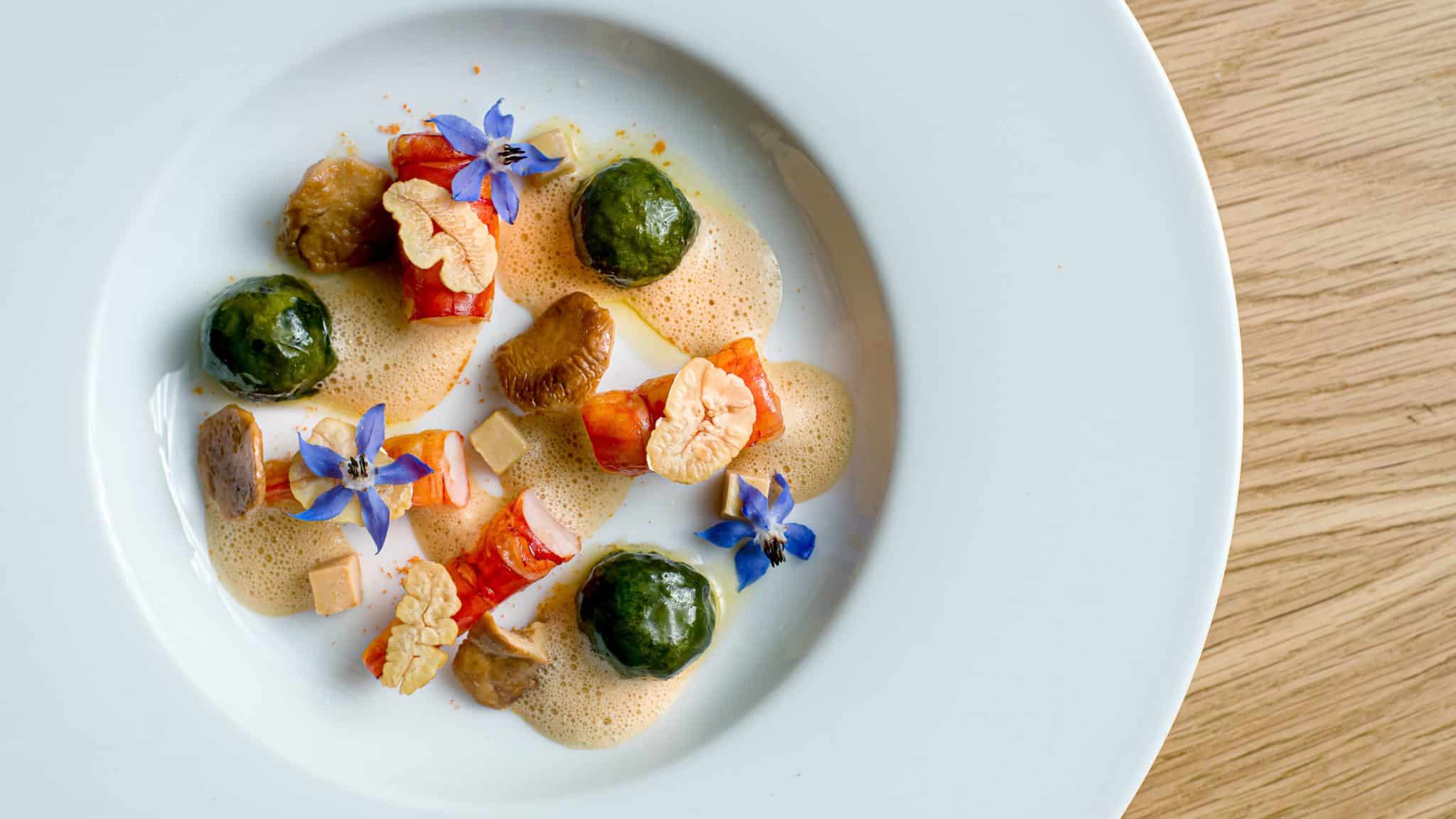 A white plate with artfully arranged gourmet food, including green spheres, diced vegetables, foam, mushrooms, edible flowers, and garnishes sits on a wooden table—an aesthetic presentation worthy of Crimson Design Group’s signature style.