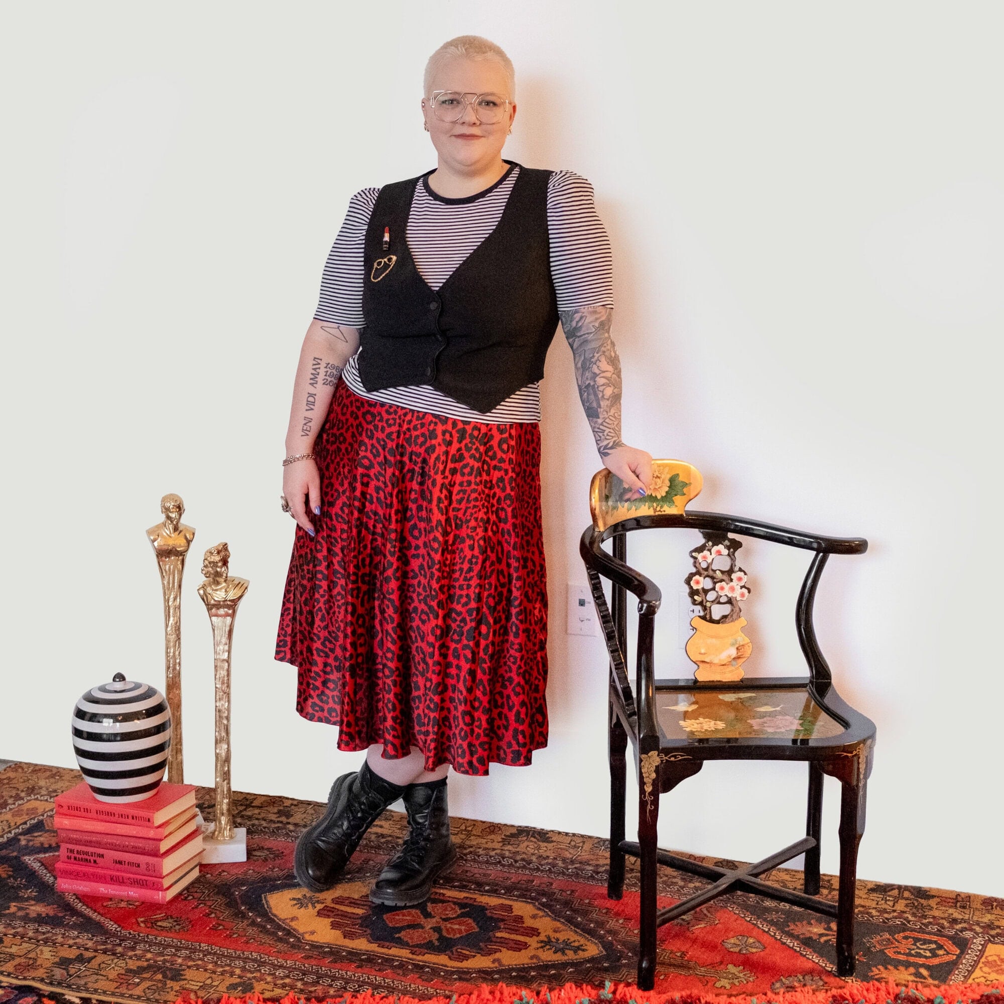 A person with short hair and tattoos stands in a striped shirt, black vest, and red leopard-print skirt, resting a hand on an ornate black chair.