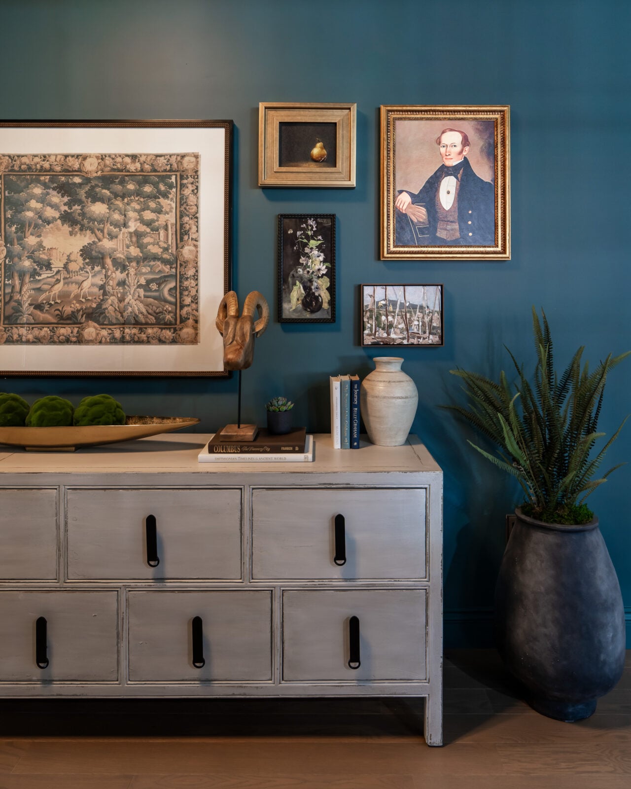 A blue wall with framed art, a white dresser with books and decor, and a tall plant in a dark pot.