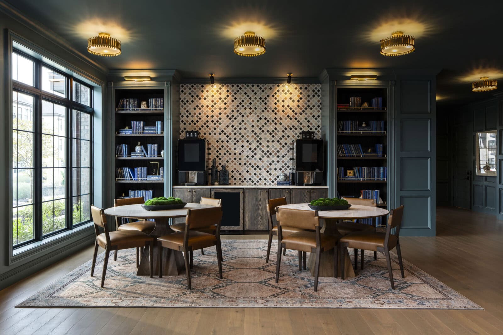 Modern lounge with two round tables, leather chairs, built-in bookshelves, patterned tile wall, and large window letting in natural light.