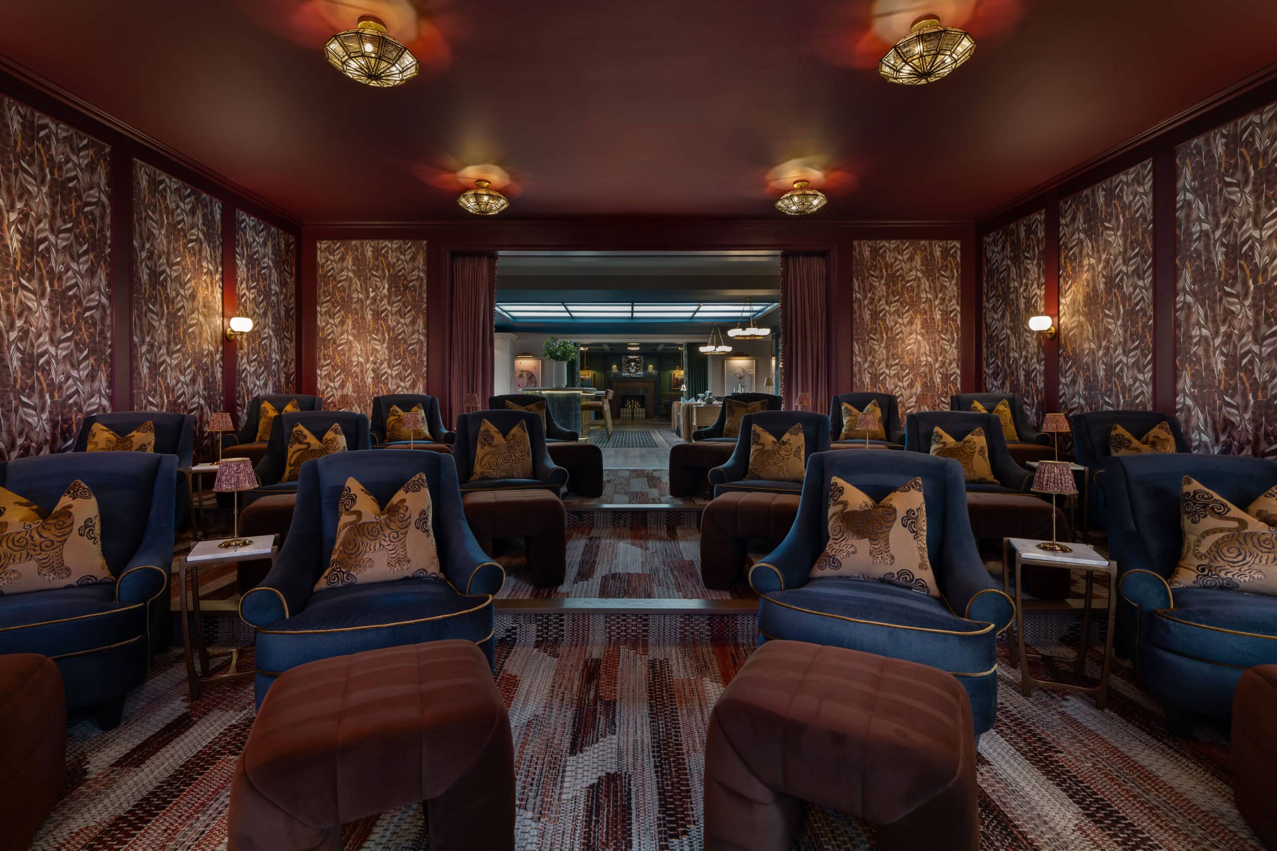 A luxurious home theater room inspired by Retail Design in Scottsdale, Arizona, featuring patterned wallpaper, rows of blue armchairs with brown pillows, matching ottomans, small side tables, and dim lighting.