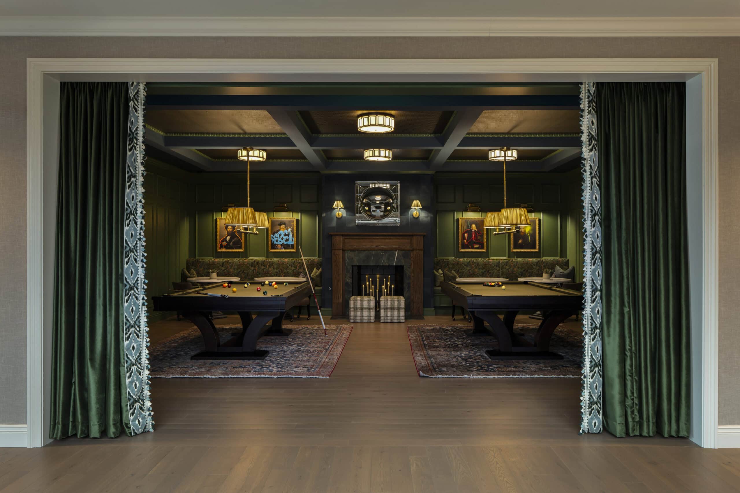 Room with green walls, two pool tables on patterned rugs, green velvet drapes, a central fireplace, and framed art on the walls. Overhead lights and lamps provide illumination.