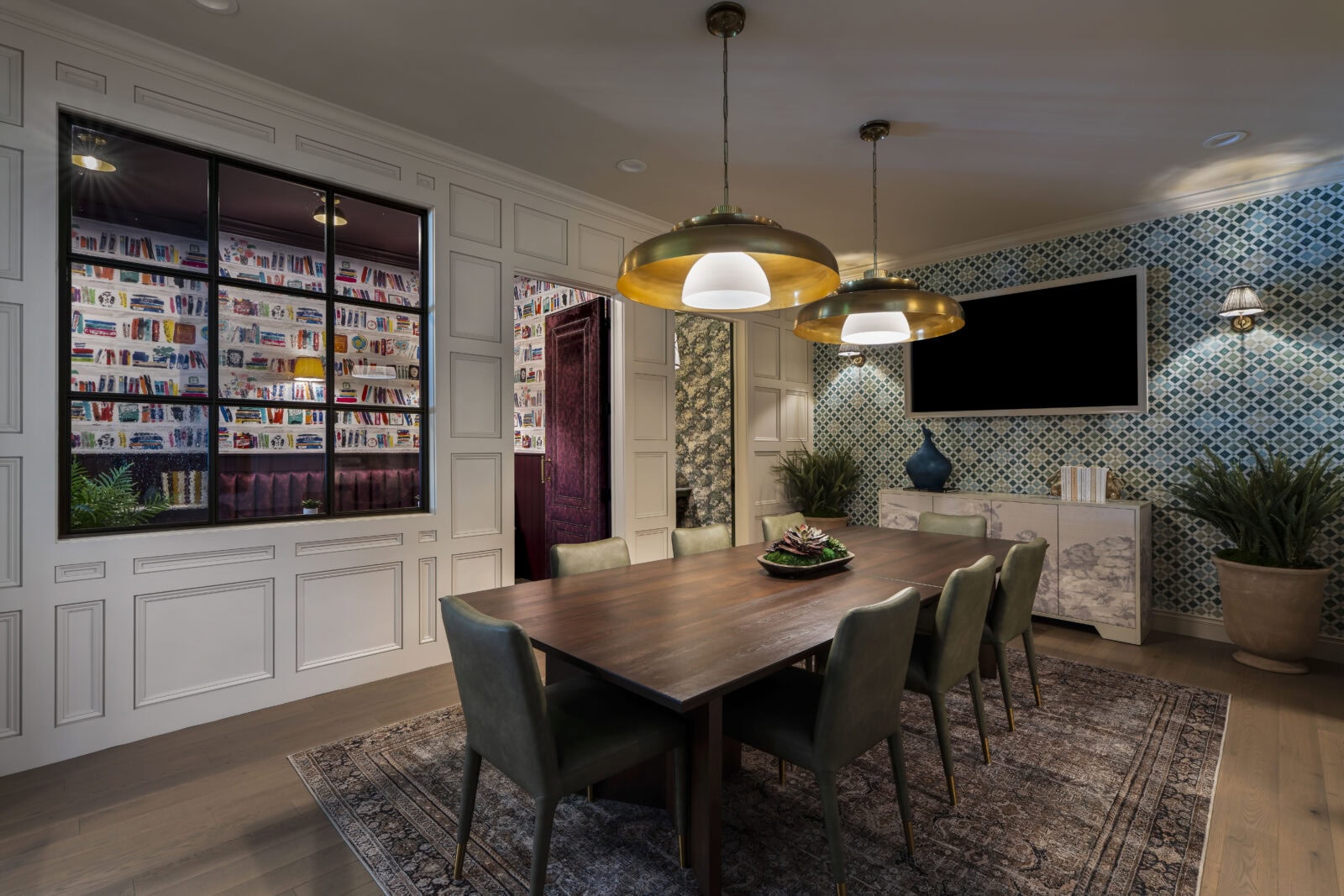 Modern dining room with a wooden table, six green chairs, three pendant lights, a wall-mounted TV, patterned wallpaper, and a window showing a colorful library.