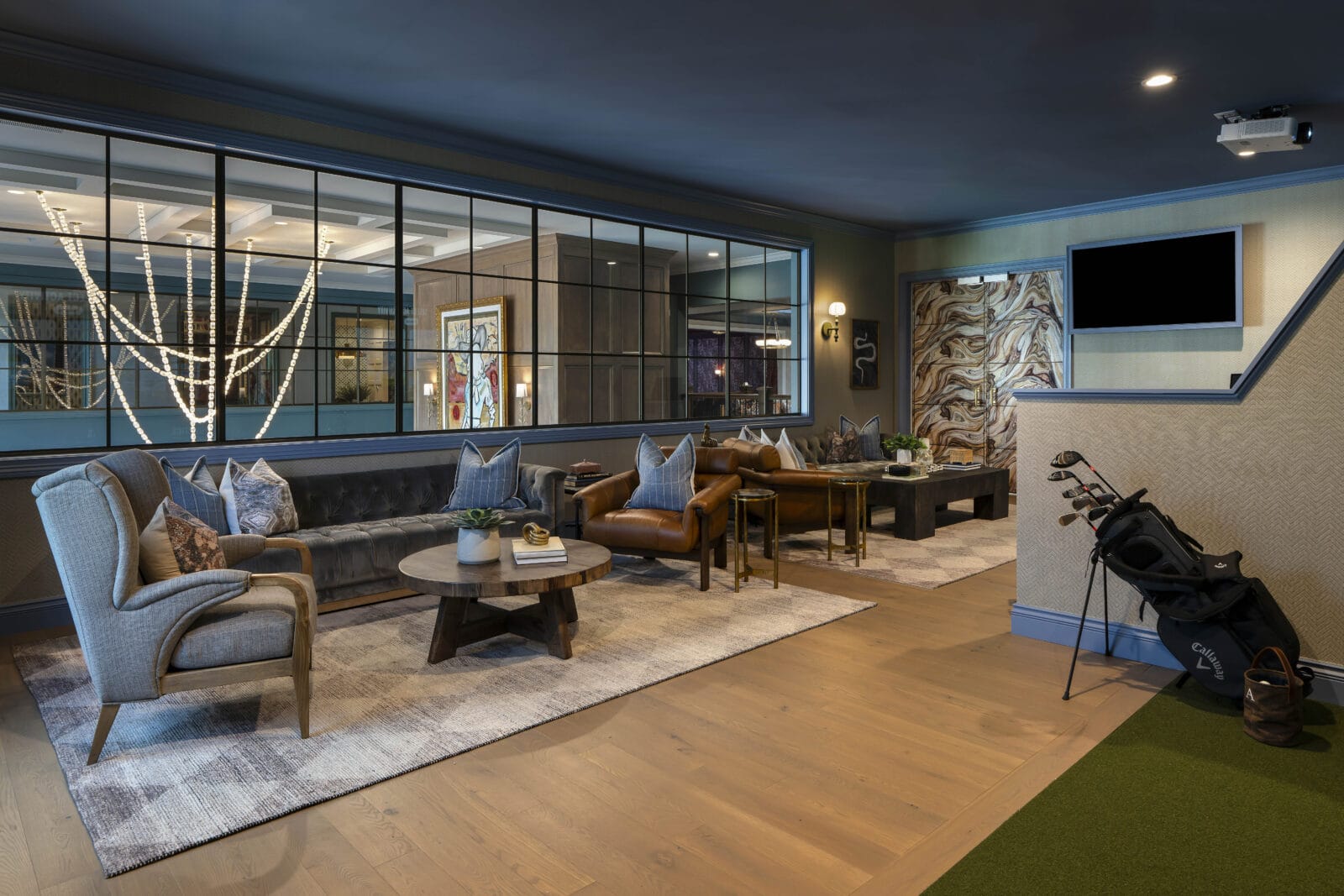 Modern lounge with leather and fabric seating, coffee tables, wall art, large window, golf bag, and putting green on wooden floors. Neutral tones and accent lighting create a cozy atmosphere.