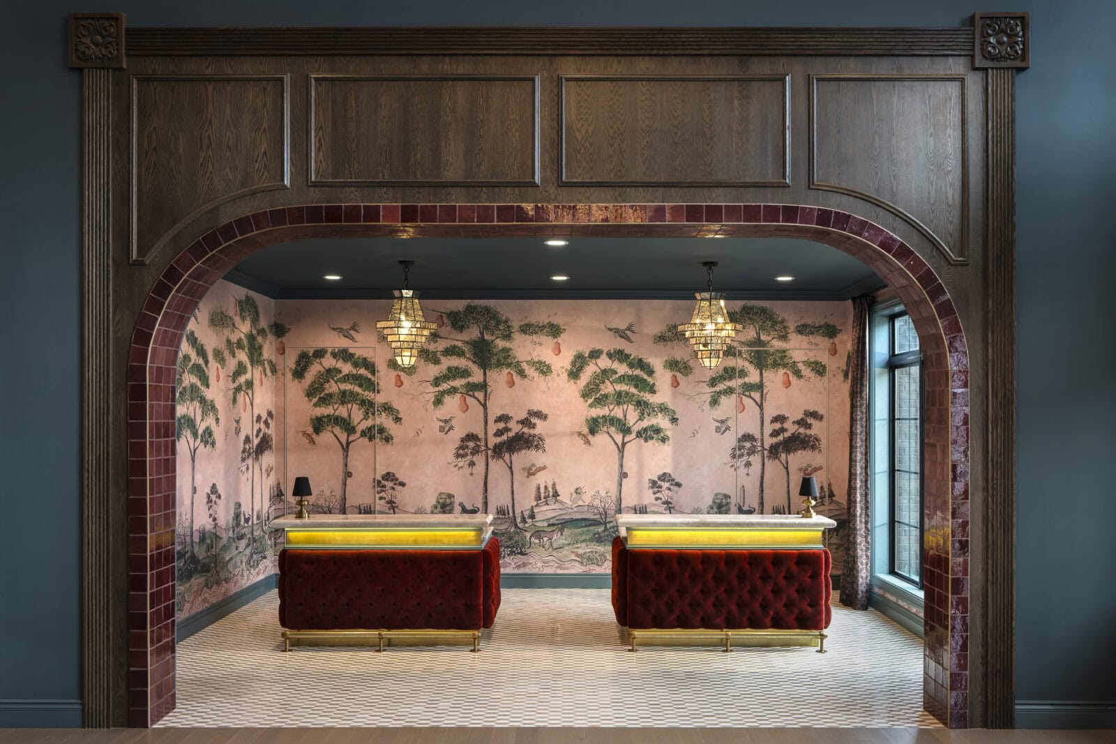 Two red velvet reception desks stand under chandeliers in a room with a pink mural of trees, framed by a wooden and red-tiled archway.