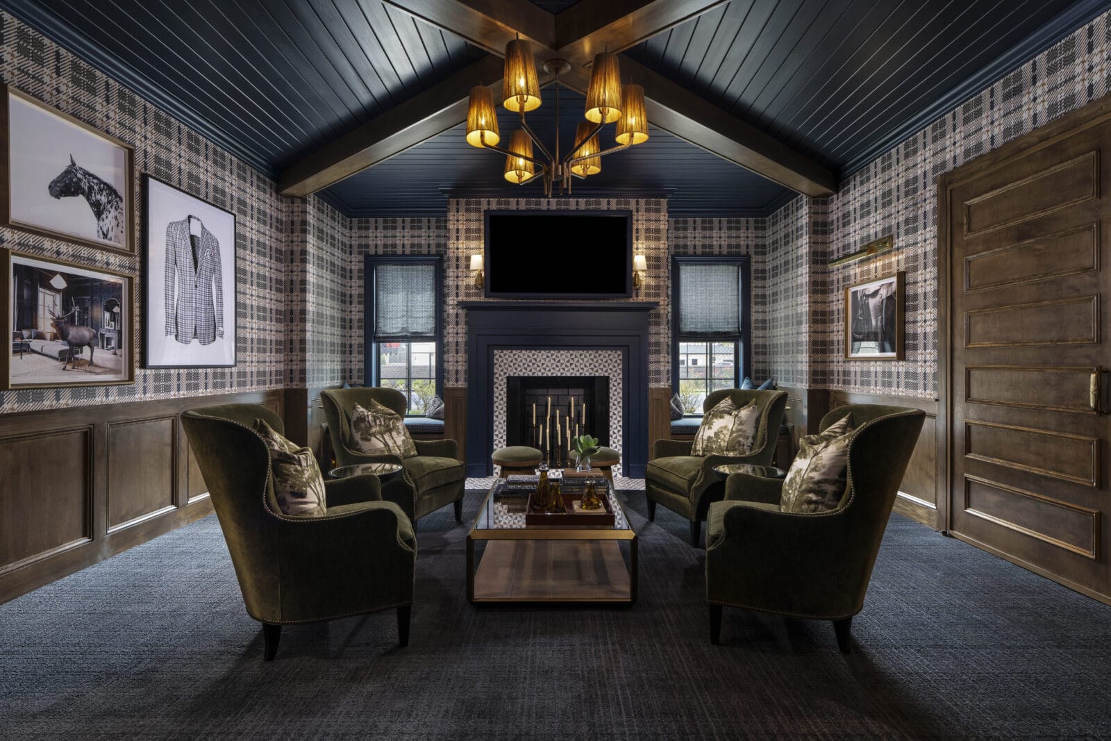 Plaid-walled lounge with six green armchairs around a wooden coffee table, fireplace, TV, chandelier, framed art, and wood-paneled door.