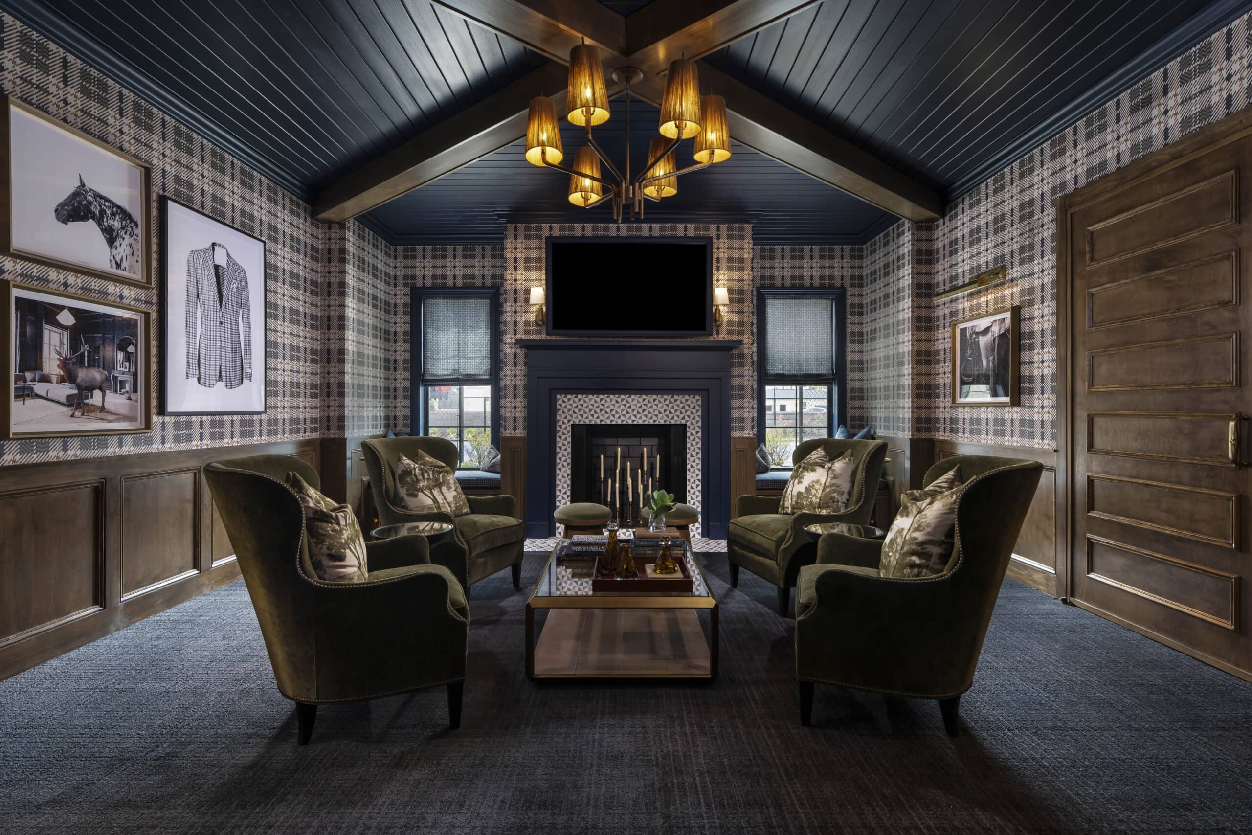 Plaid-walled lounge with six green armchairs around a wooden coffee table, fireplace, TV, chandelier, framed art, and wood-paneled door.