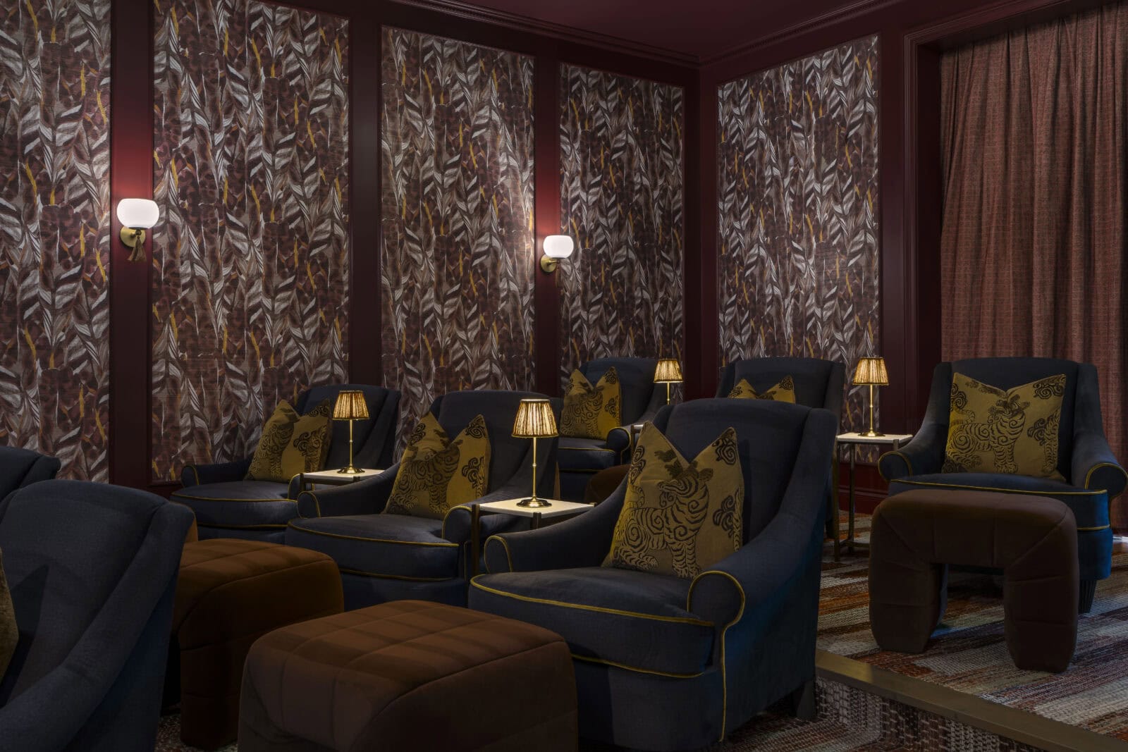 A cozy room with navy armchairs, brown ottomans, small table lamps, patterned pillows, and decorative wallpaper, arranged like a private screening or lounge area.