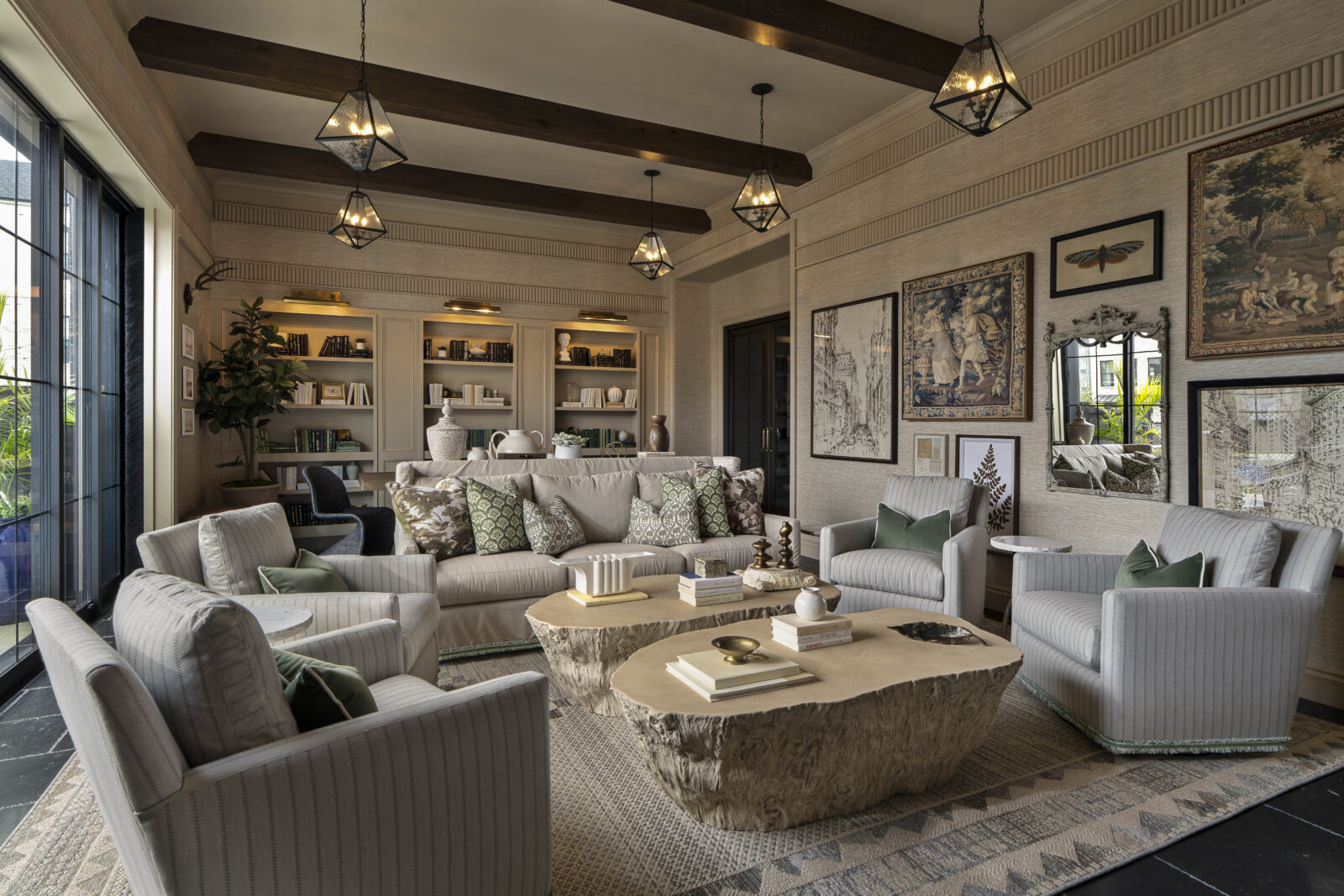 Elegant living room with neutral furniture, exposed ceiling beams, built-in shelves, framed art on walls, and two stone coffee tables on a patterned rug. Large windows provide natural light.