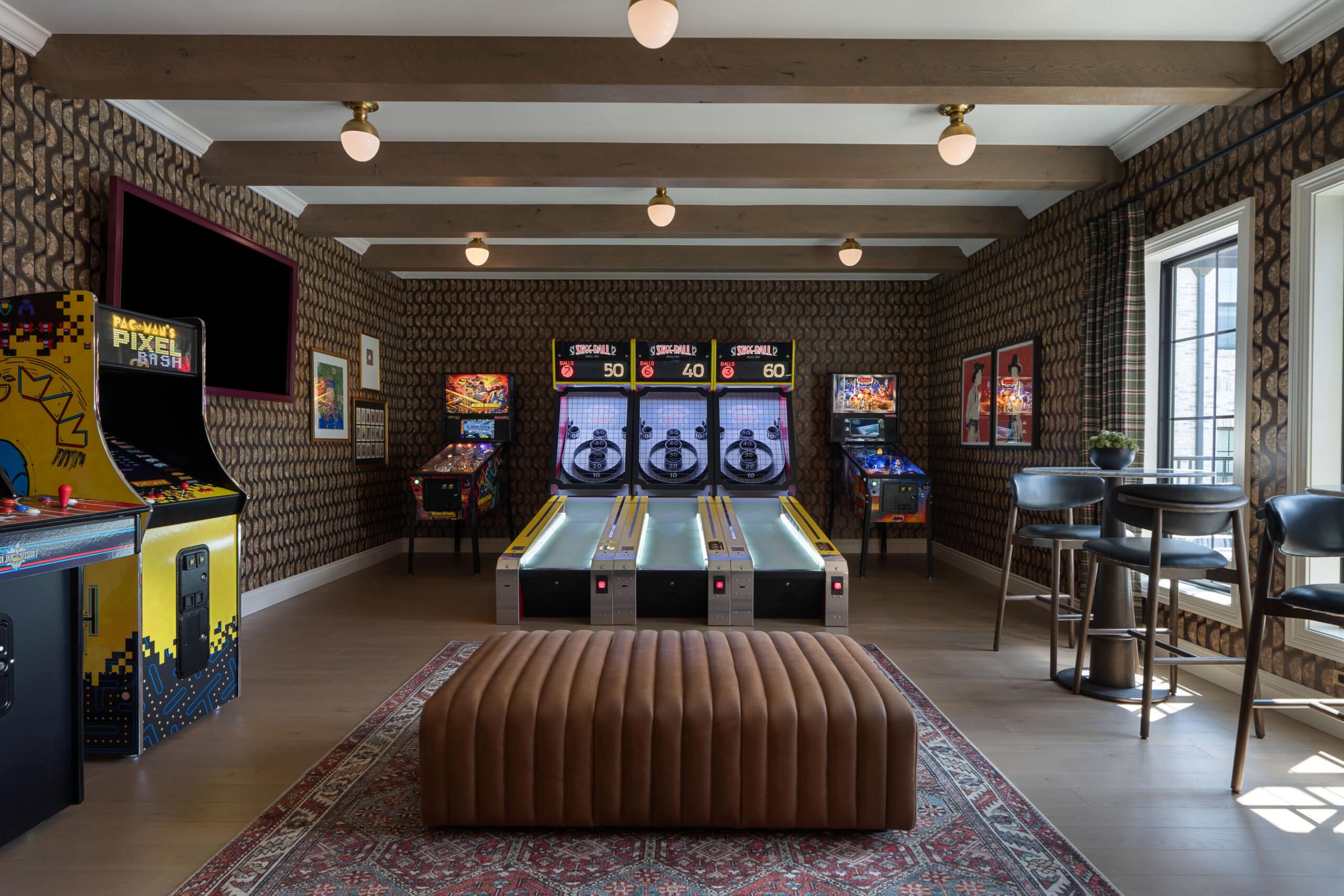 Game room with arcade machines, skee-ball lanes, pinball machines, a brown ottoman, high chairs, patterned wallpaper, and a rug on wooden flooring—an inviting space that draws inspiration from innovative Retail Design in Scottsdale Arizona.