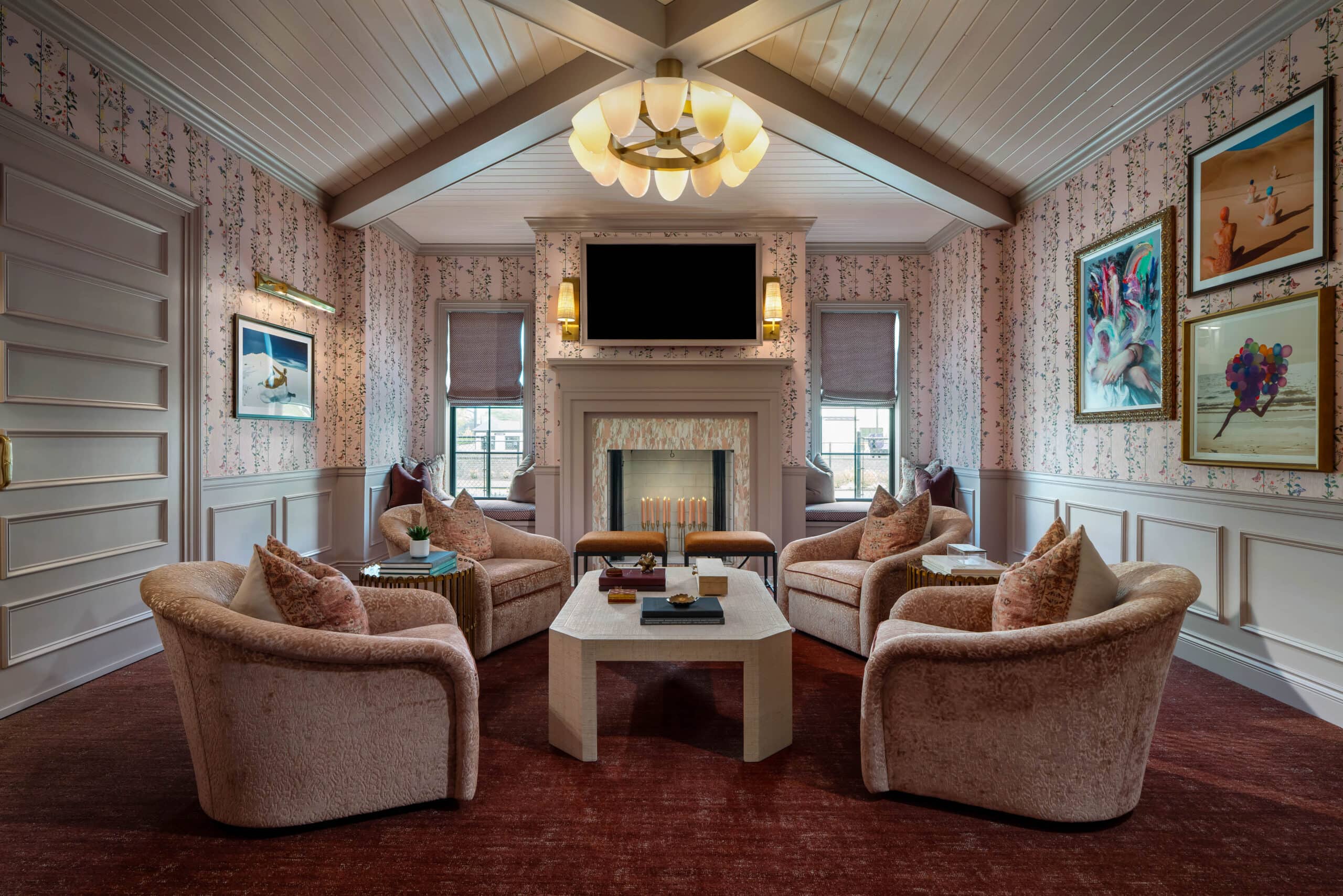 A cozy living room inspired by retail design in Scottsdale, Arizona, features four plush chairs around a square coffee table, a fireplace, wall art, floral wallpaper, and a chandelier under a vaulted ceiling.