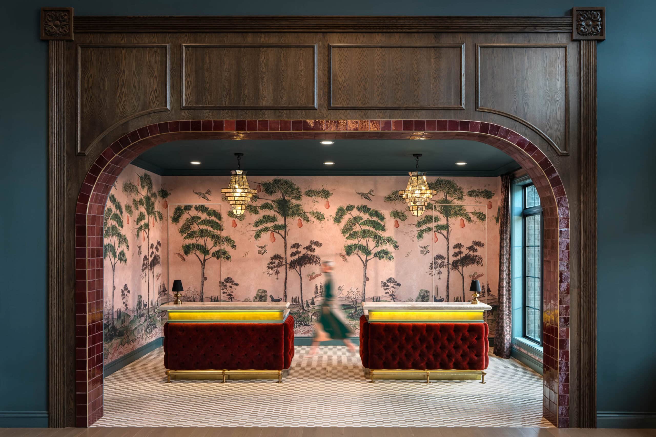 Hotel reception area with two red velvet desks, wallpaper featuring trees, chandeliers above, and a person walking past; framed by a large wooden arch with red tile accents—a striking example of retail design in Scottsdale, Arizona.