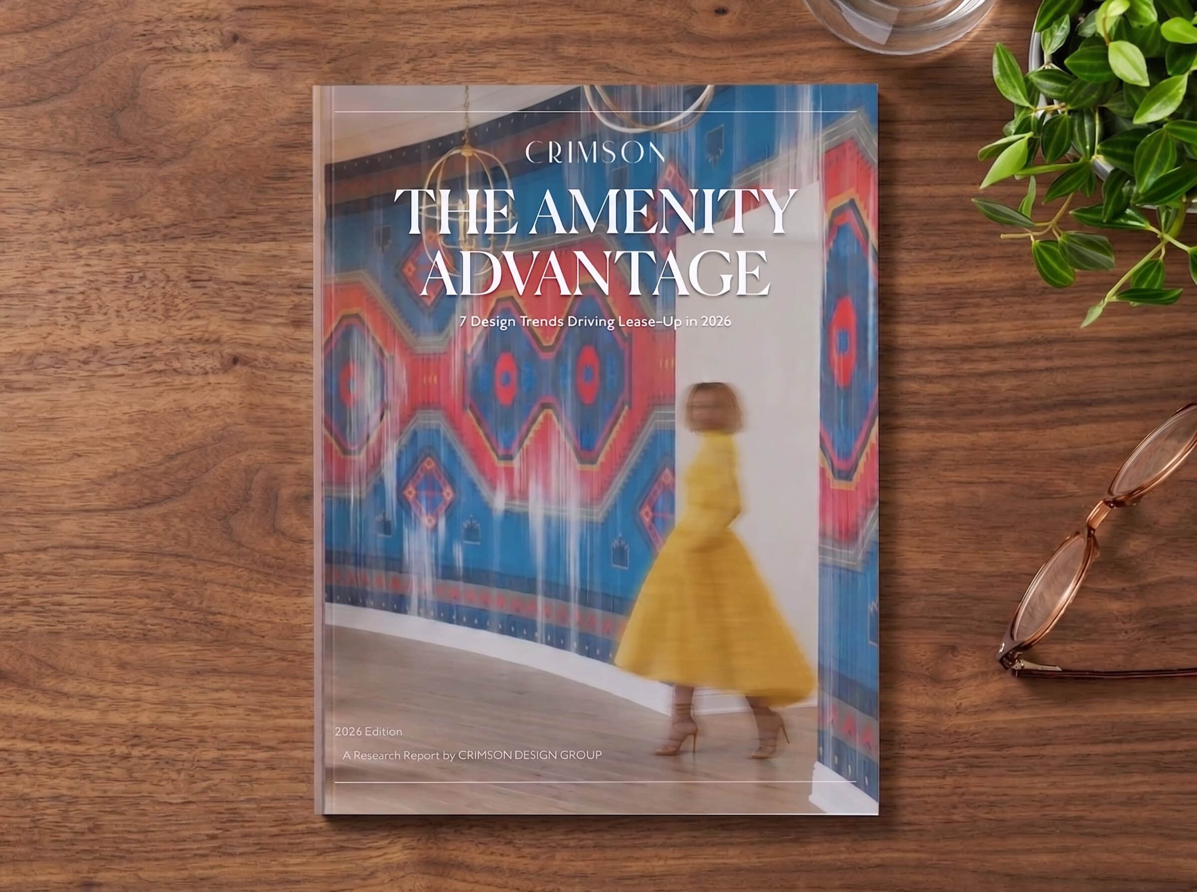 A magazine titled "The Amenity Advantage" with a blurred image of a woman in a yellow dress walking past a colorful geometric mural, placed on a wooden table next to glasses and a plant.