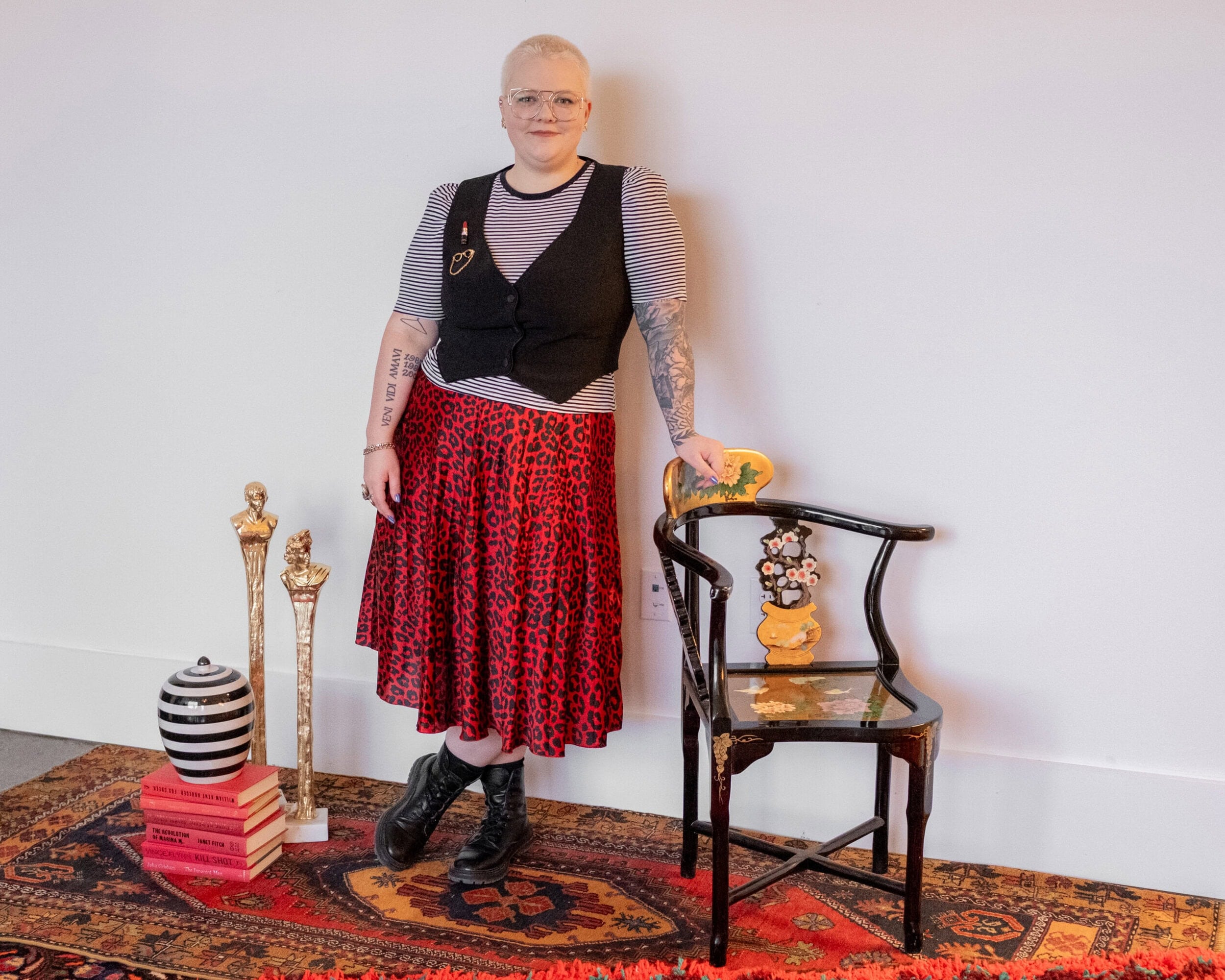 A person with short hair, glasses, and tattoos stands by a chair on a patterned rug. Decorative items and stacked books are arranged nearby against a plain white wall.