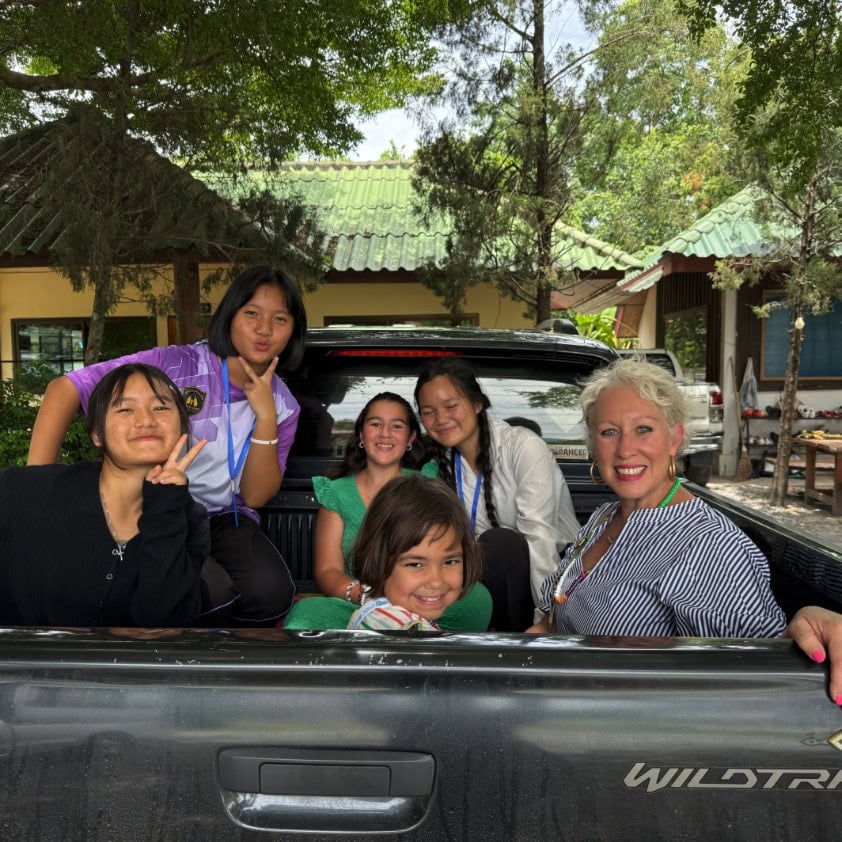 Six people, including five young girls and one older woman, sit and pose for a group photo in the back of a black pickup truck outside a building surrounded by trees.