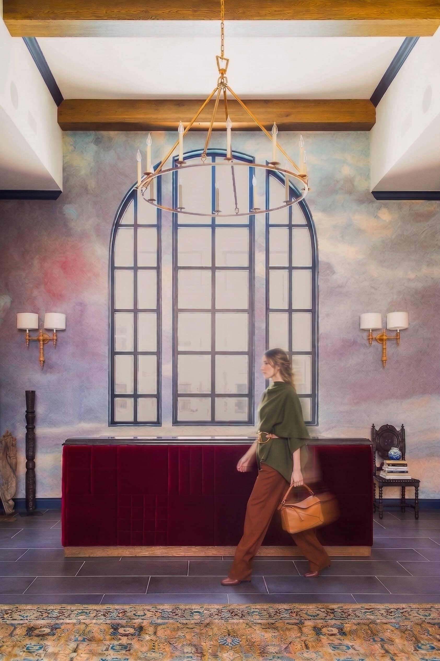 A person walks past a red velvet reception desk in a lobby with a large arched window, gold chandelier, wall sconces, and a decorative rug.