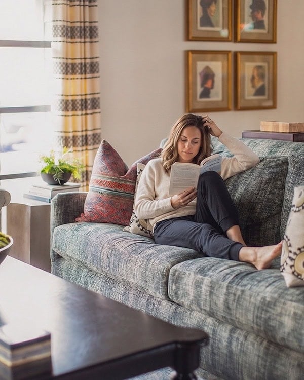 A woman sits on a blue sofa reading a book in a well-lit living room with patterned pillows, framed art, and a large window.